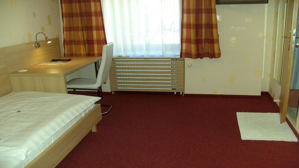 A simply furnished hotel room with a single bed, desk and chair. Red carpet and checkered curtains.