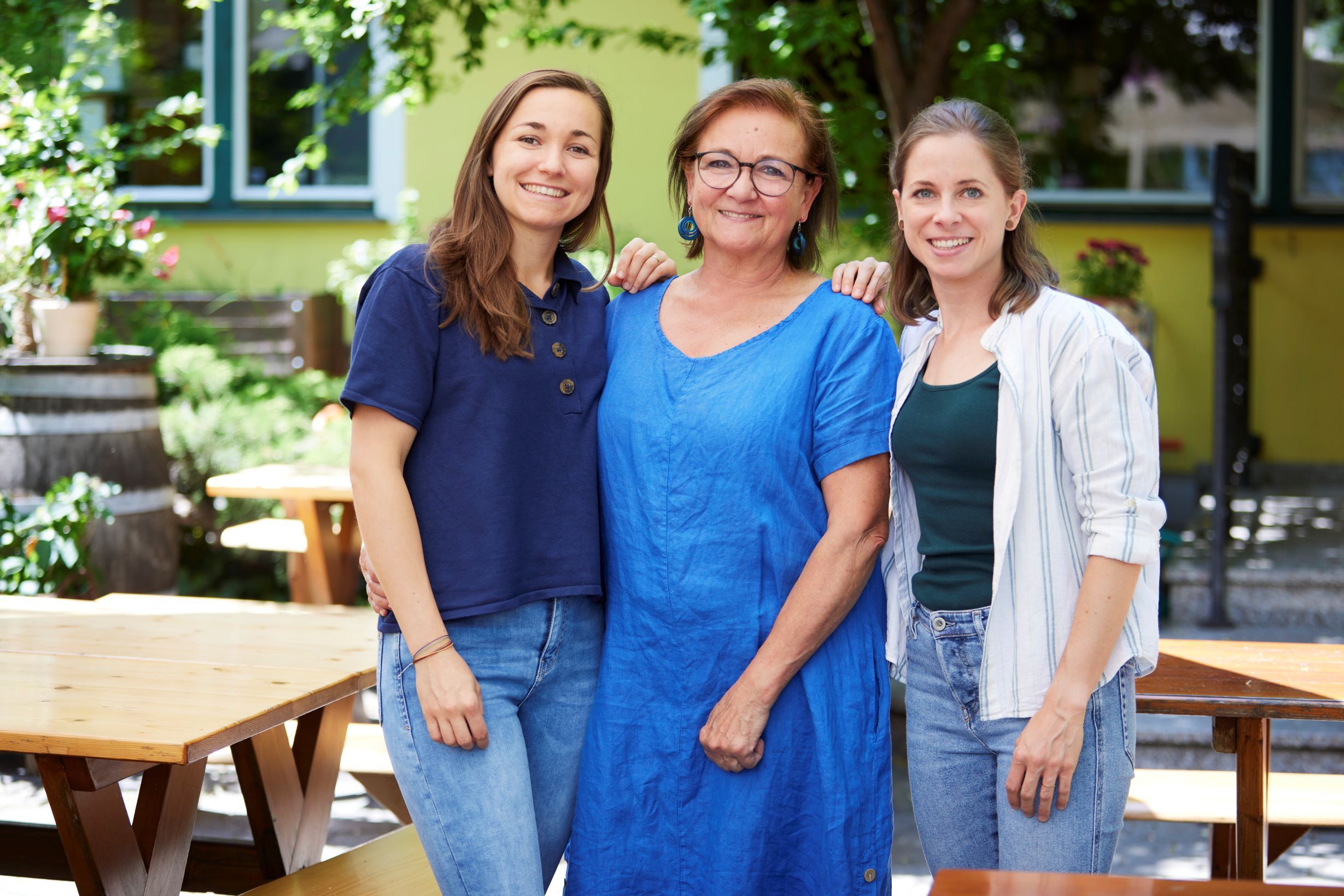 Three women stand smiling in a garden with wooden tables and plants.
