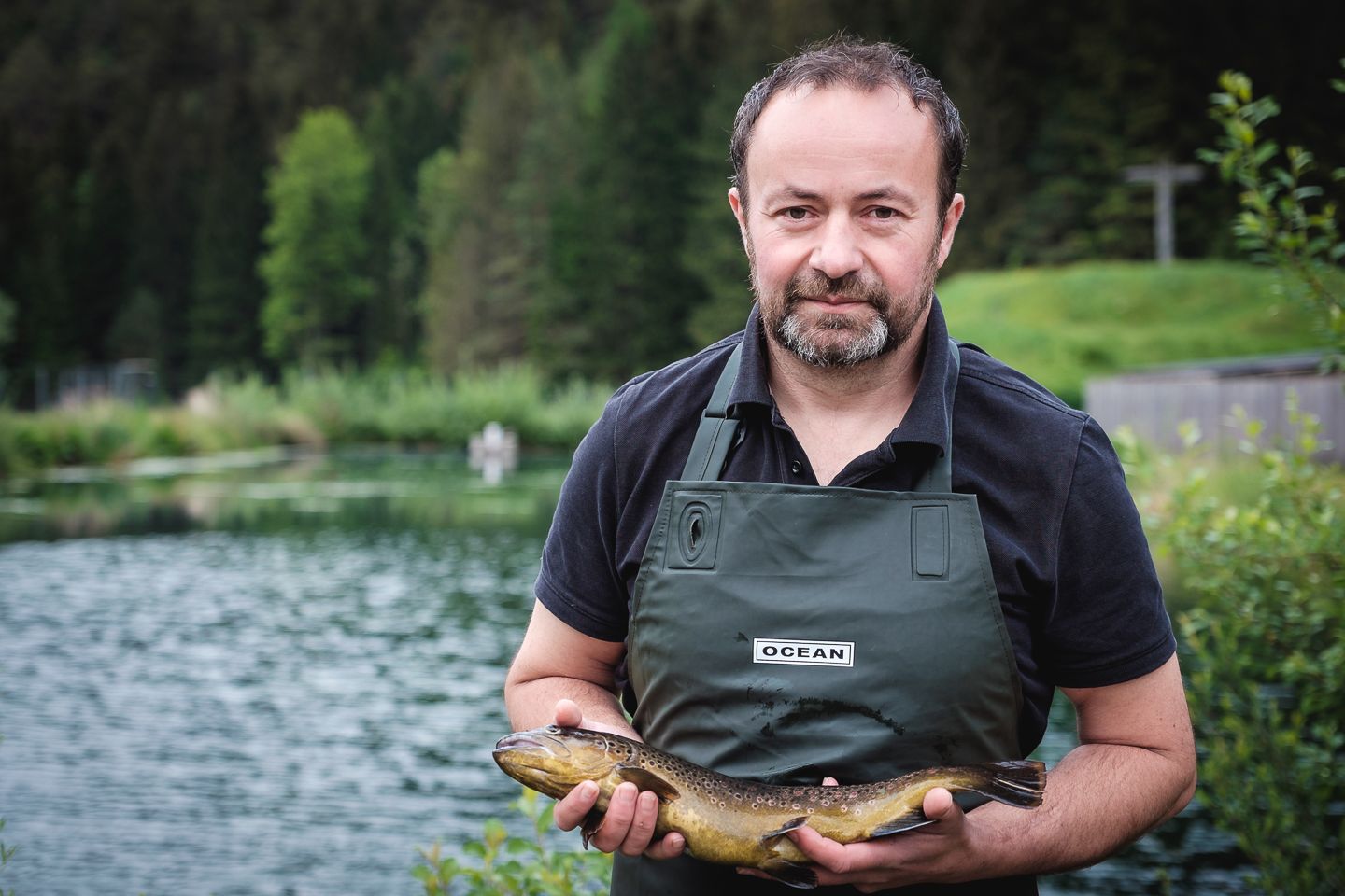 A man in a green apron holds a fish in front of a pond.