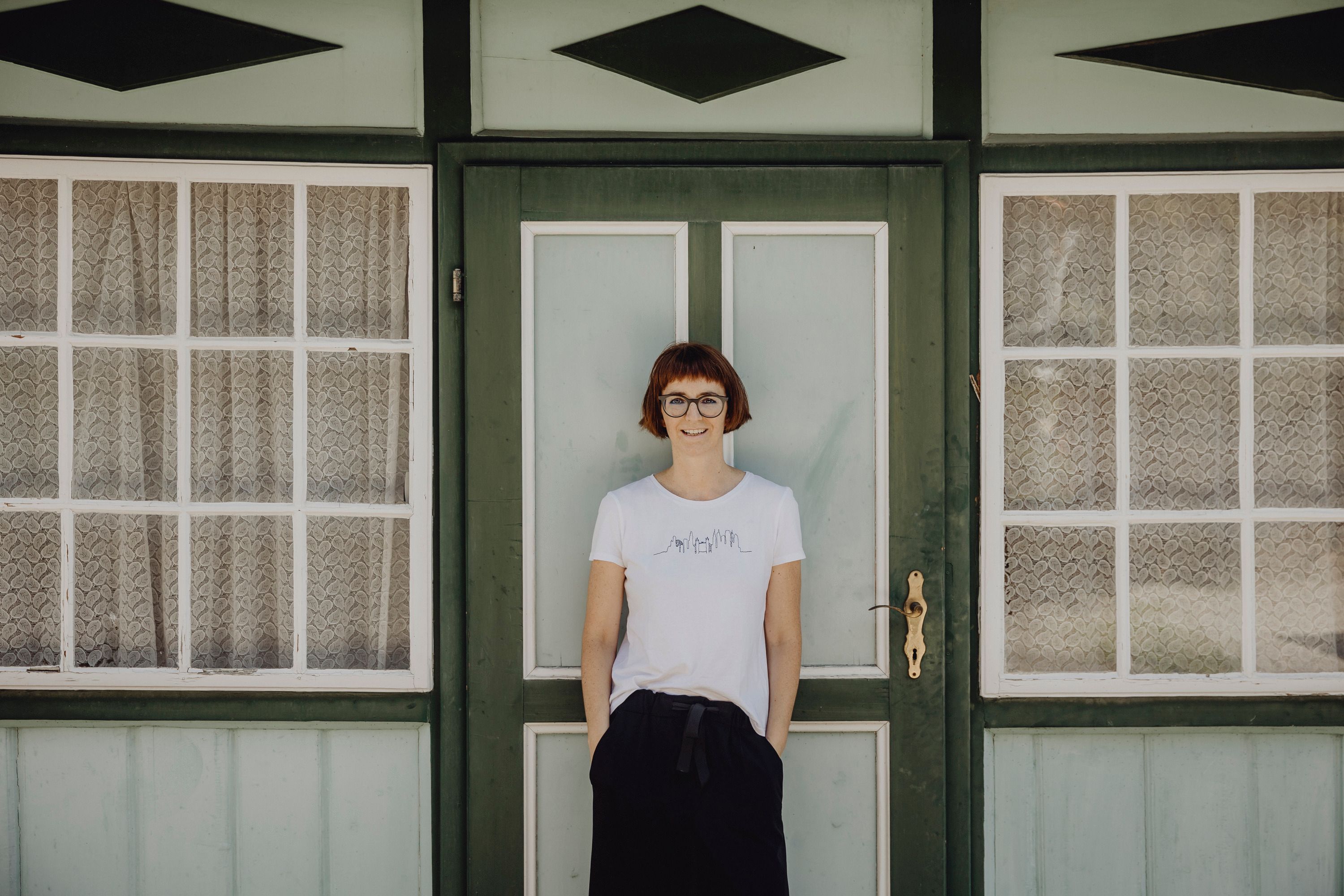 A woman stands in front of a green door with windows and lace curtains.