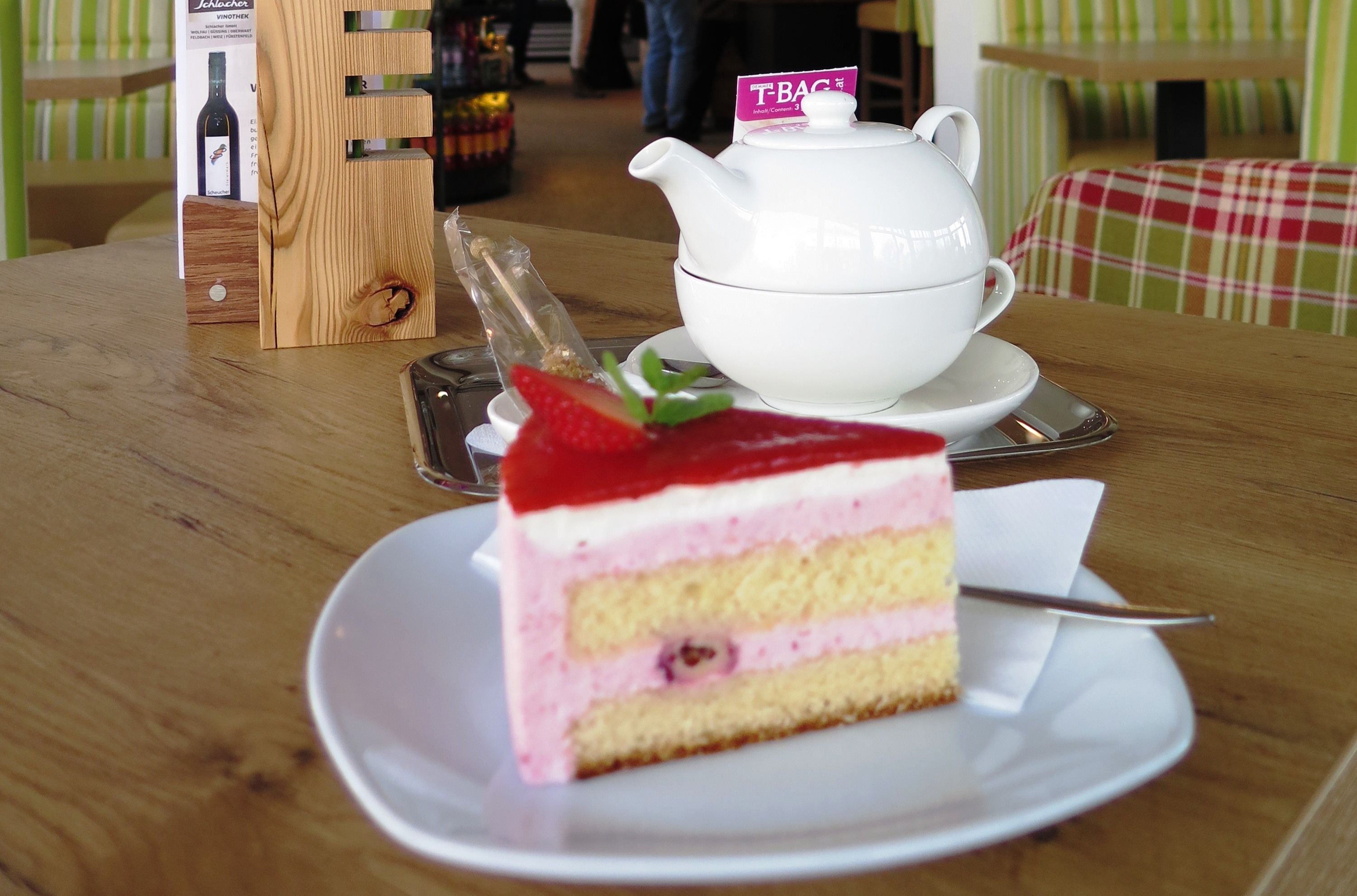 A slice of strawberry cake on a plate with a teapot in the background on a wooden table.