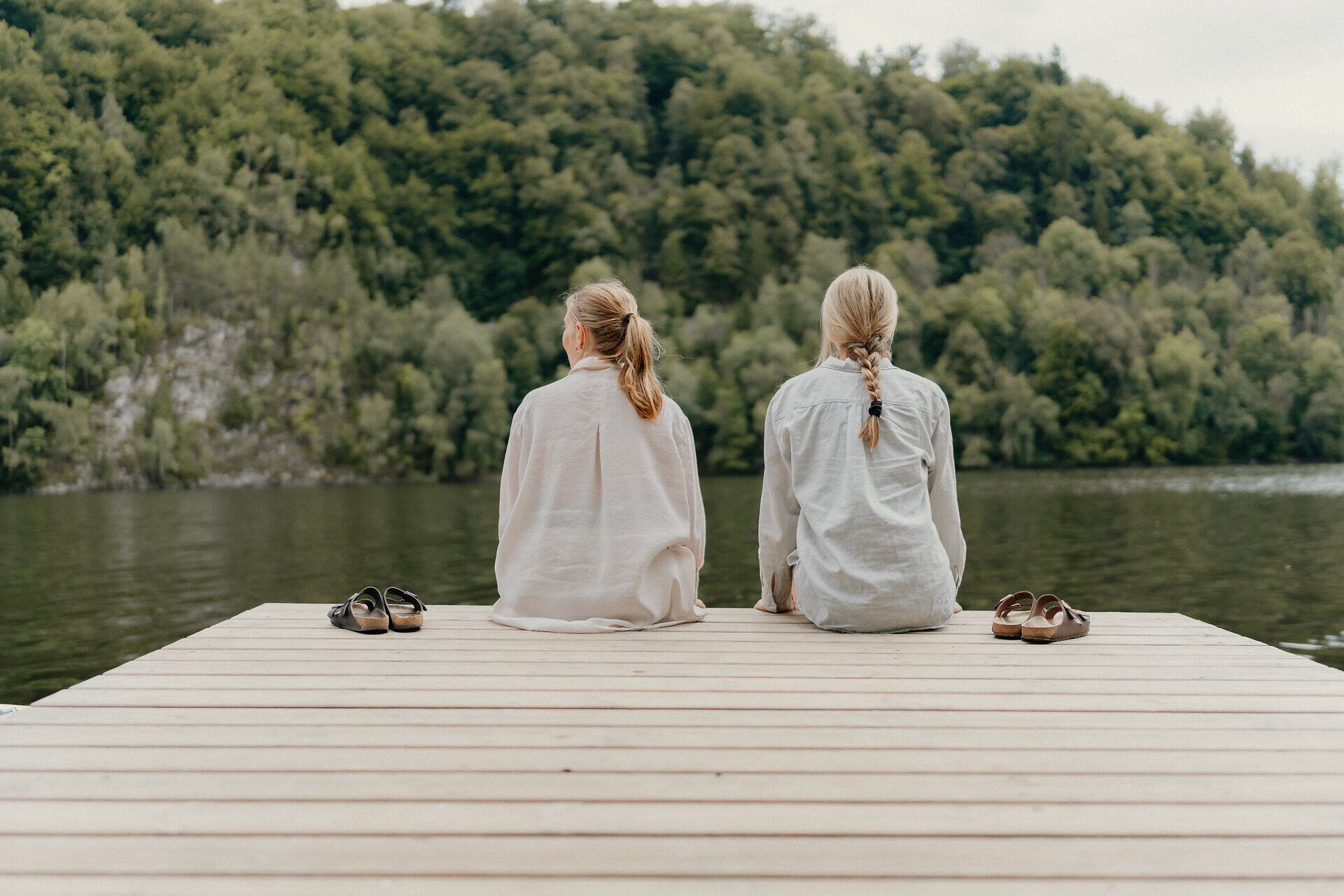 Zwei Personen sitzen barfuß auf einem Holzsteg am Stausee Dobra im Waldviertel; ihre Schuhe stehen neben ihnen, gegenüber liegt ein bewaldetes Ufer.