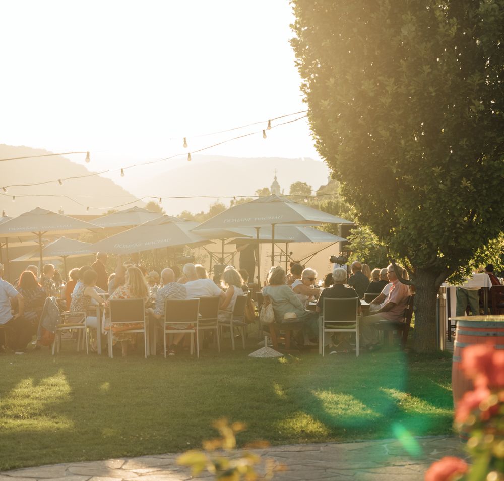 In der warmen Abendsonne versammeln sich Menschen in einem malerischen Garten, um die kulinarischen Köstlichkeiten der Region zu genießen. Die sanften Klänge von Musik und das Lachen der Gäste schaffen eine einladende Atmosphäre, während die umliegenden Weinreben in goldenem Licht erstrahlen.