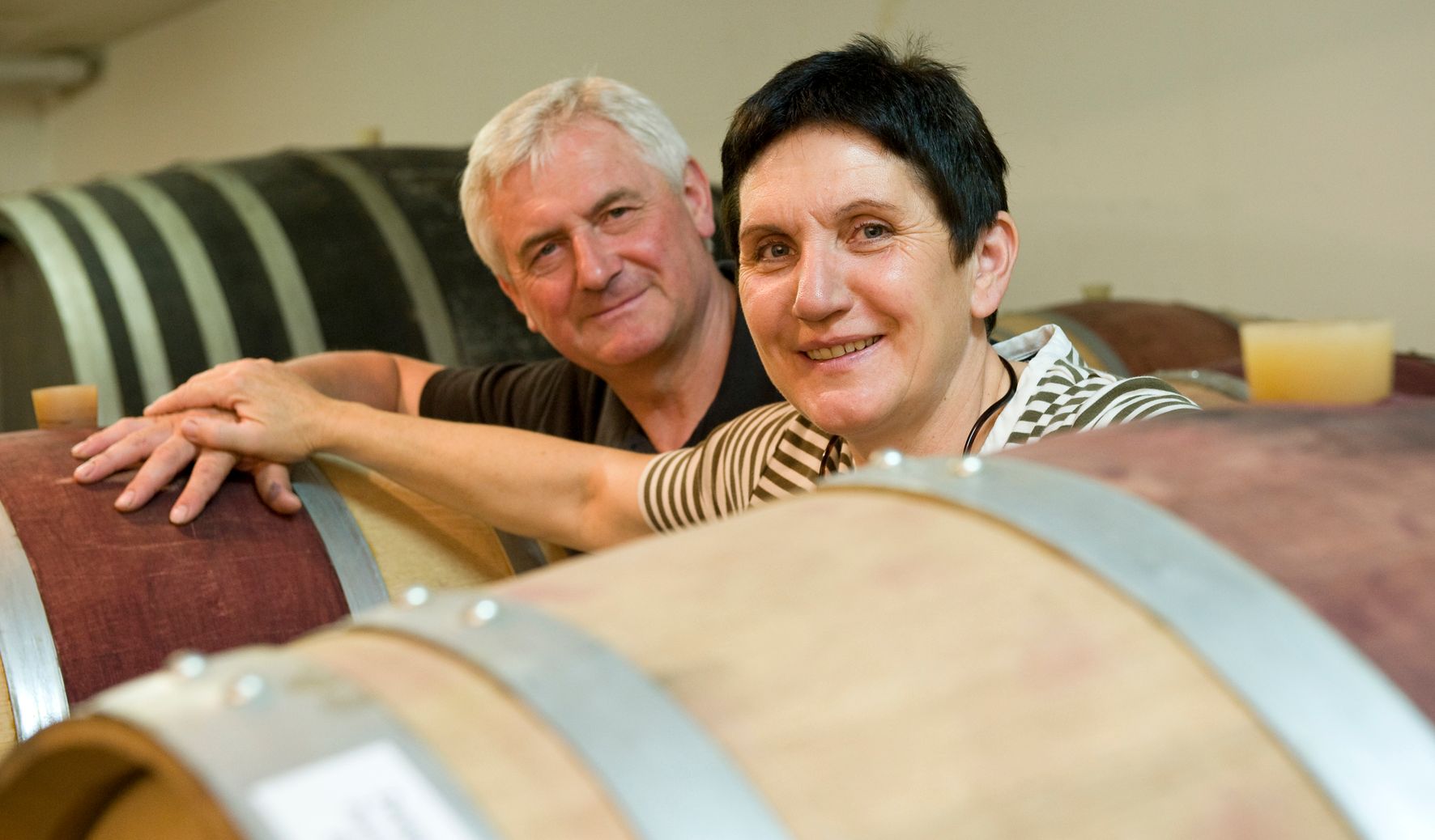 Two people smile next to wooden barrels in a winery.
