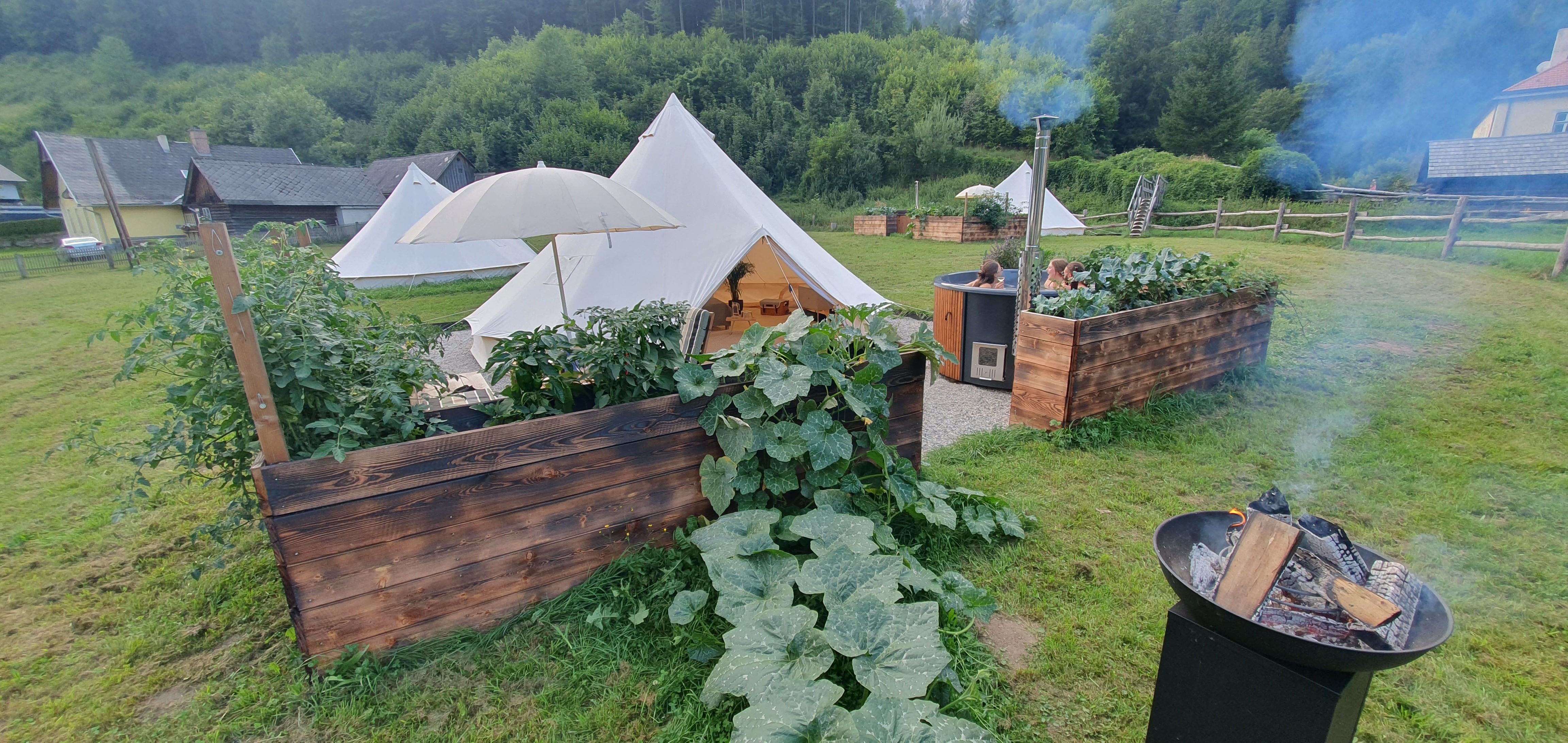 Glamping campsite with fire pit and hot tub in green surroundings.
