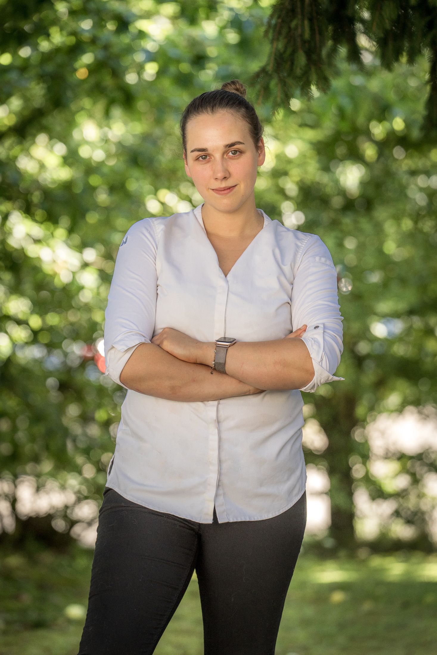 A woman in a white shirt stands with her arms folded in front of a blurred green background.