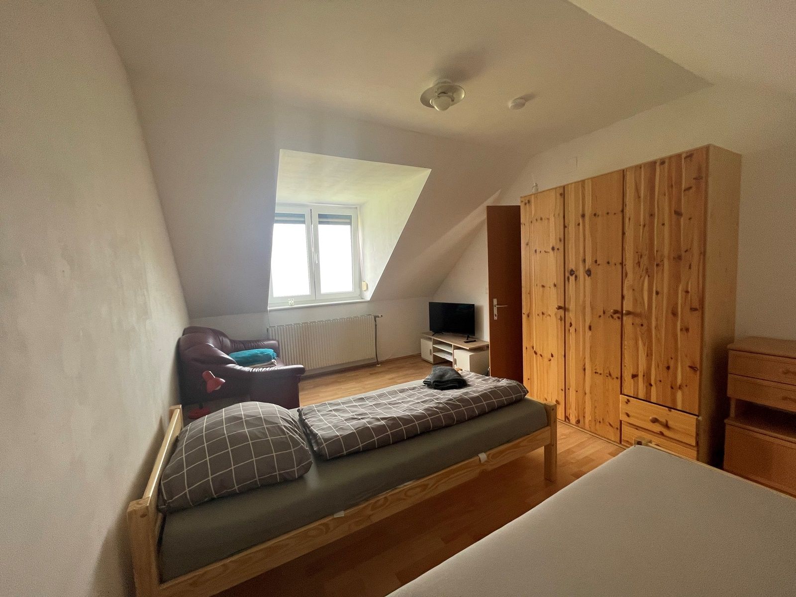 A simply furnished bedroom with two beds, a closet, a TV and an armchair under a sloping roof.