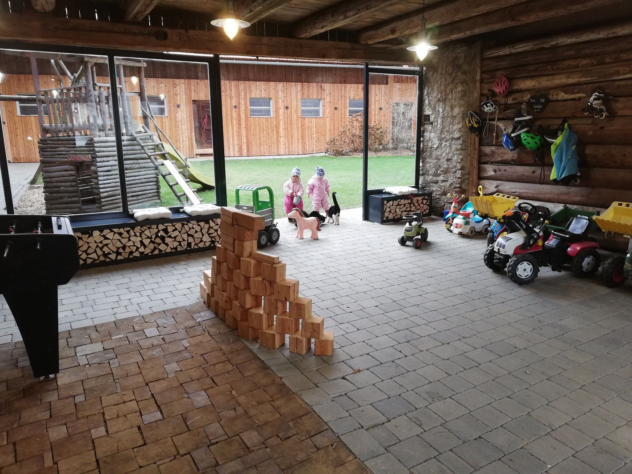 Interior view of a play barn with toy cars, wooden blocks and two children in winter clothing.