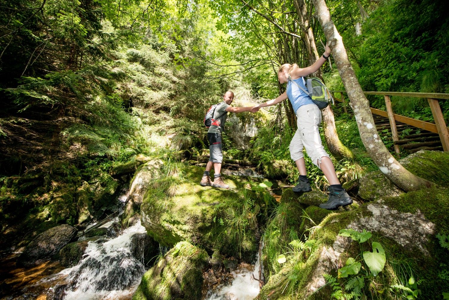 Two hikers help each other cross a stream in a green forest.