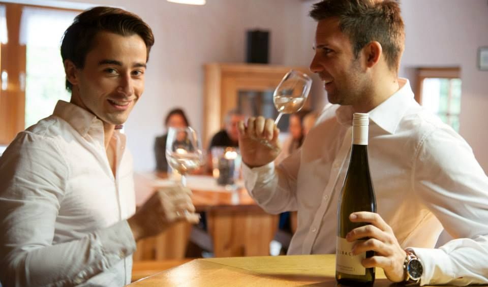 Two men in white shirts drink wine in a cozy room.