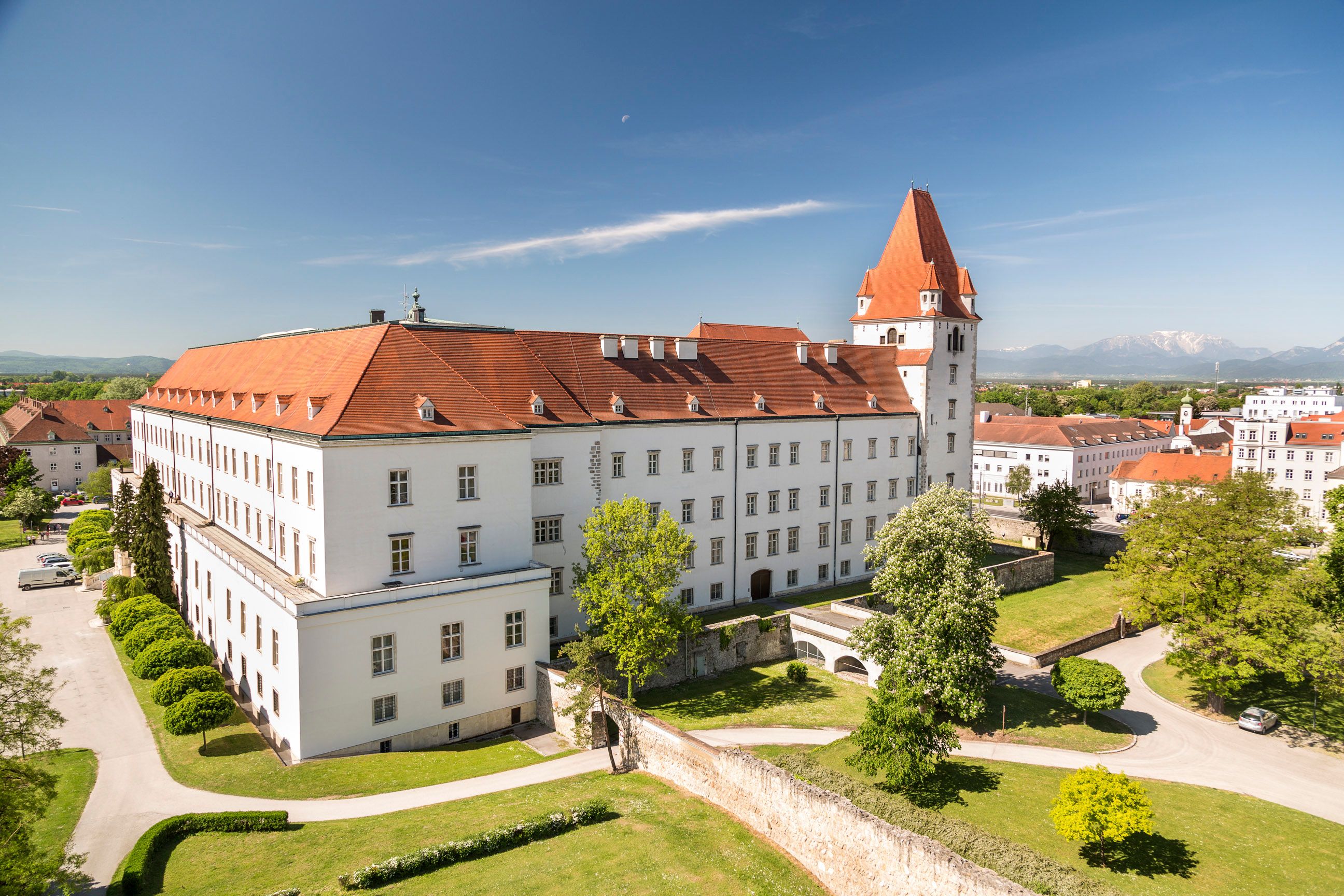 View of a historic military academy with a red roof and tower, surrounded by green trees and lawns, mountains in the background.