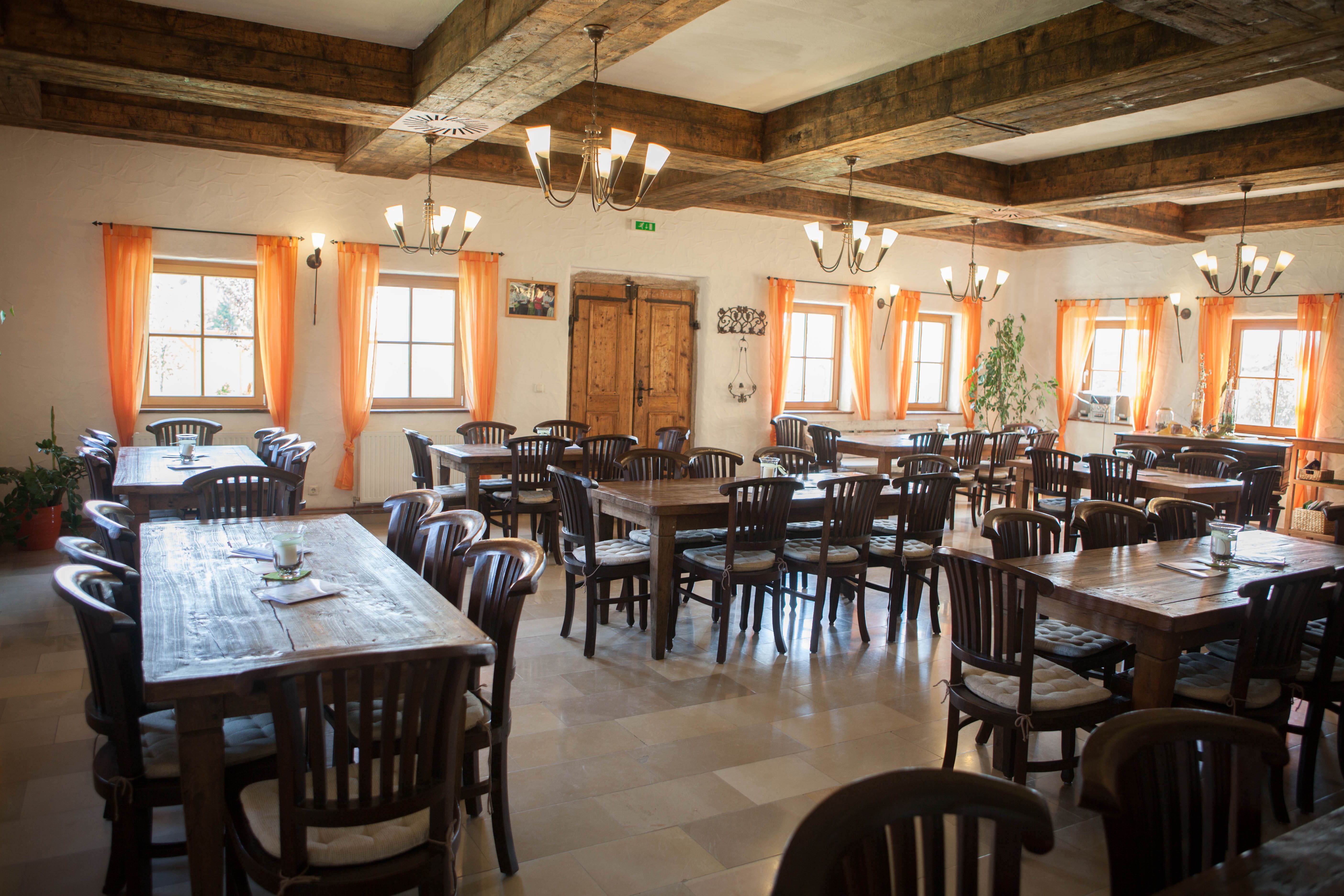 A rustic dining room with wooden tables and chairs, orange curtains and a wooden beamed ceiling.