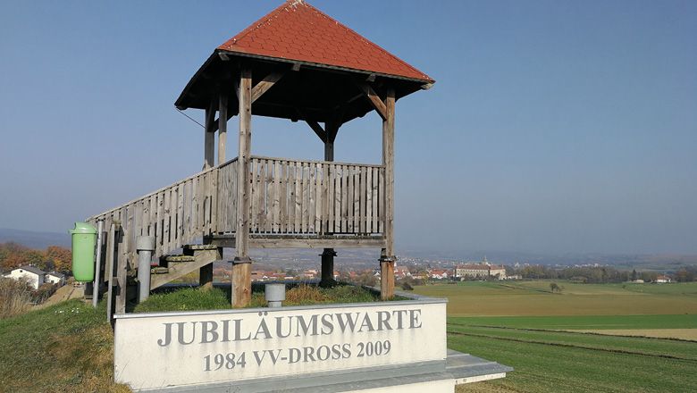Wooden lookout with a red roof on a hill, surrounded by fields and a village in the background.