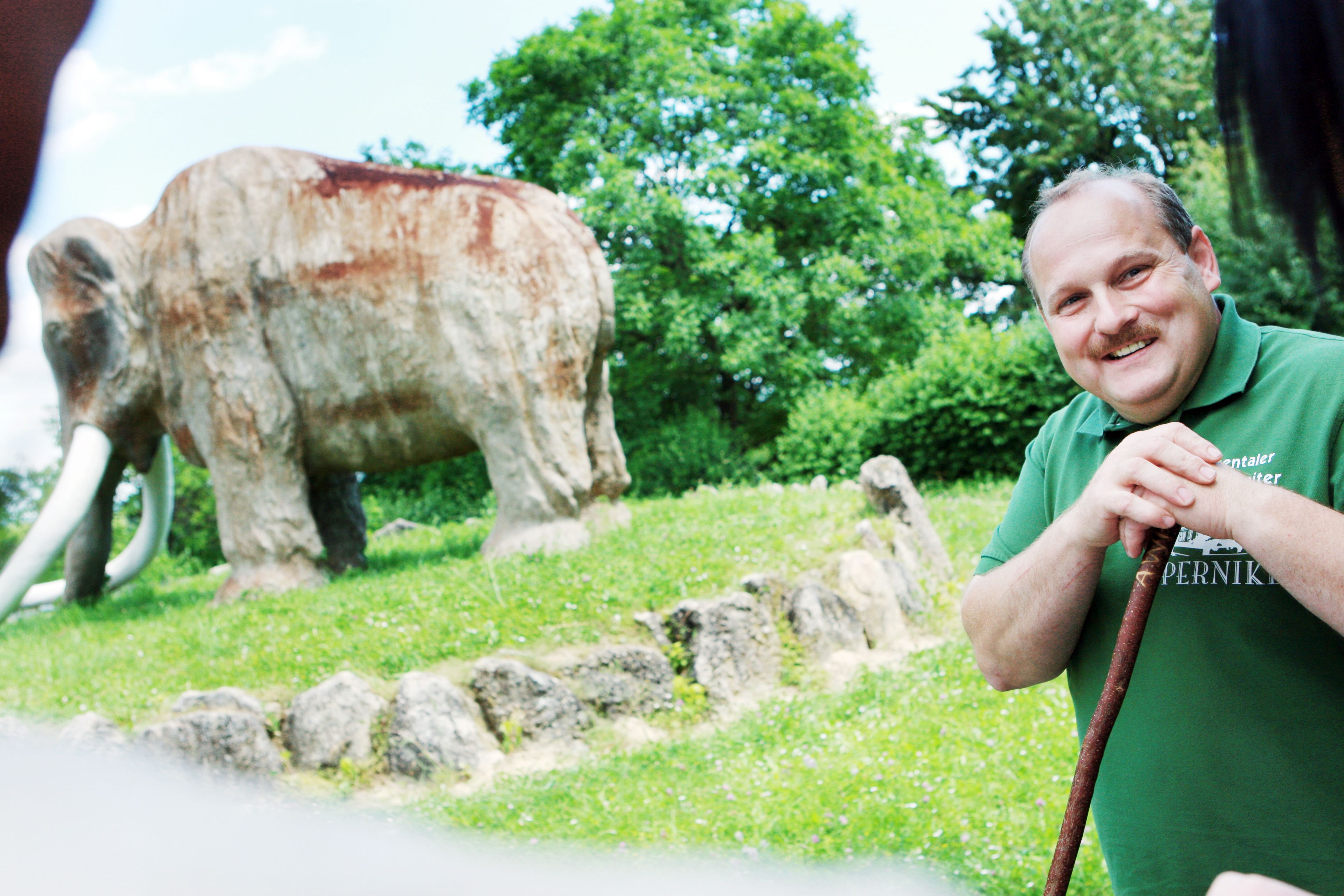 A man smiles in front of a large mammoth statue outdoors.