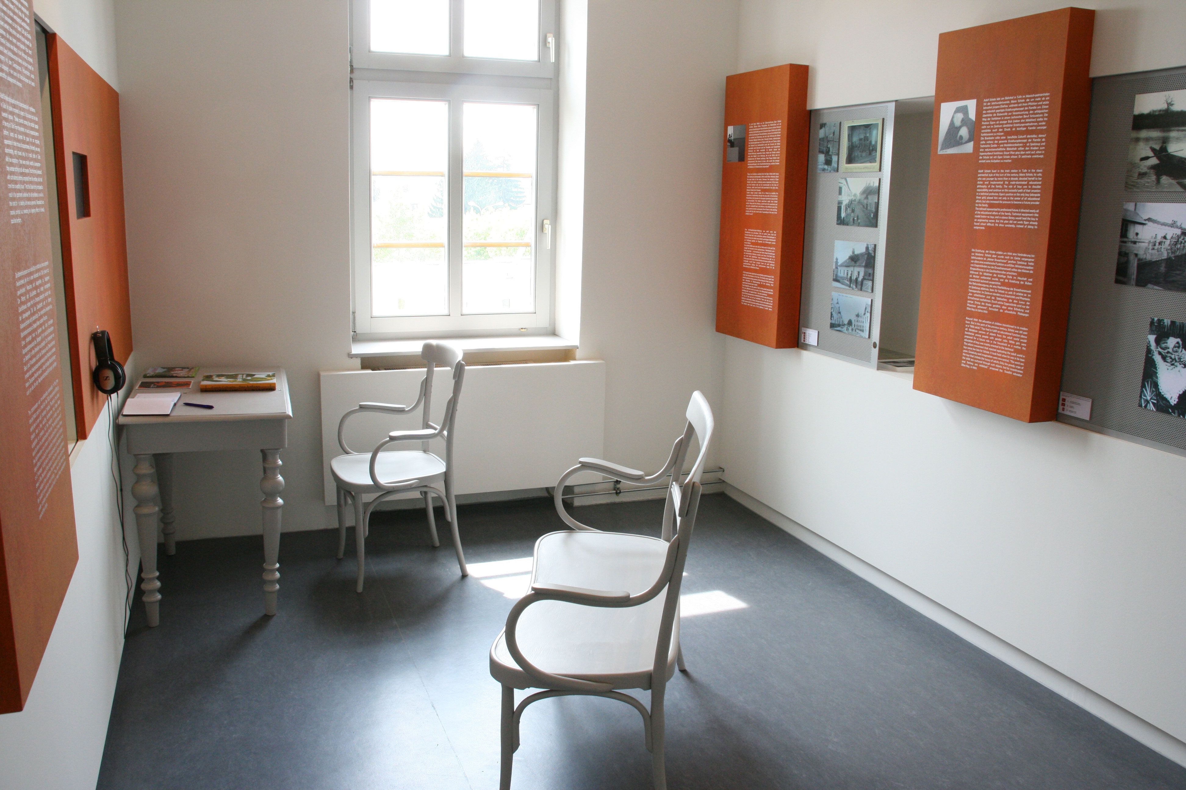 Interior view of a museum room with white chairs, a table and information boards on the walls.