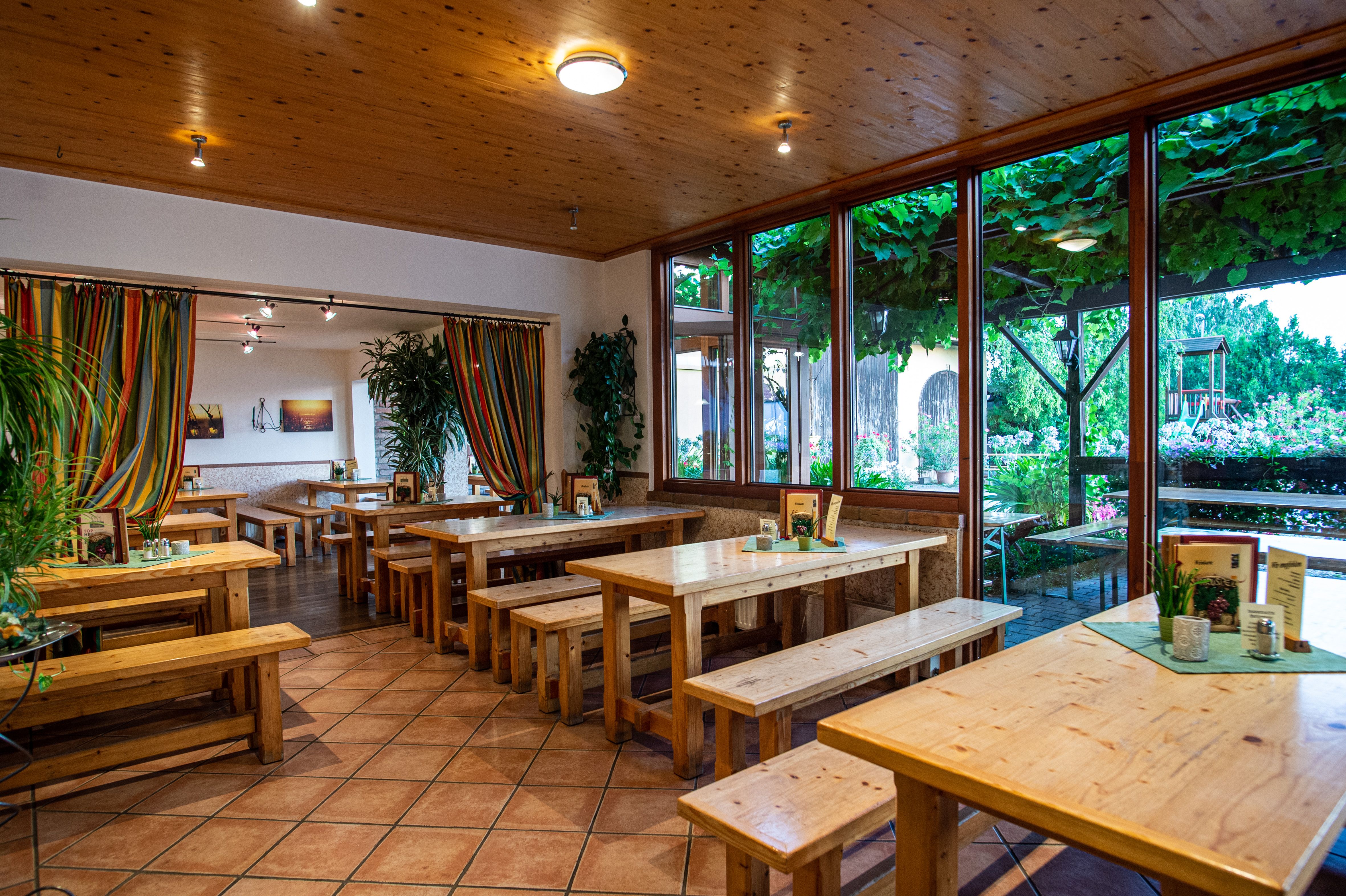 Interior view of a wine tavern with wooden tables and benches, colorful curtains and large windows overlooking a leafy outdoor area.