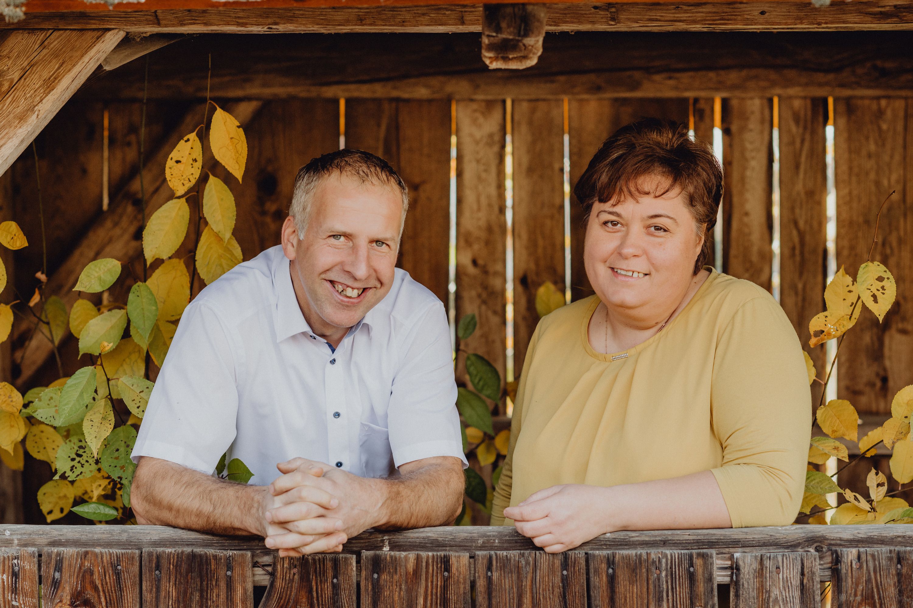 A man and a woman stand smiling by a wooden fence with autumn leaves in the background.