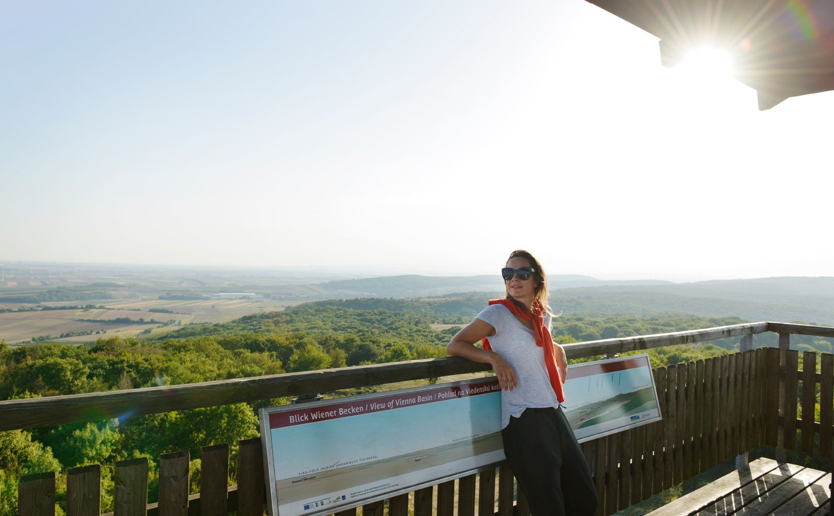 Woman with sunglasses leaning against a railing on a lookout tower with a view of a vast landscape.
