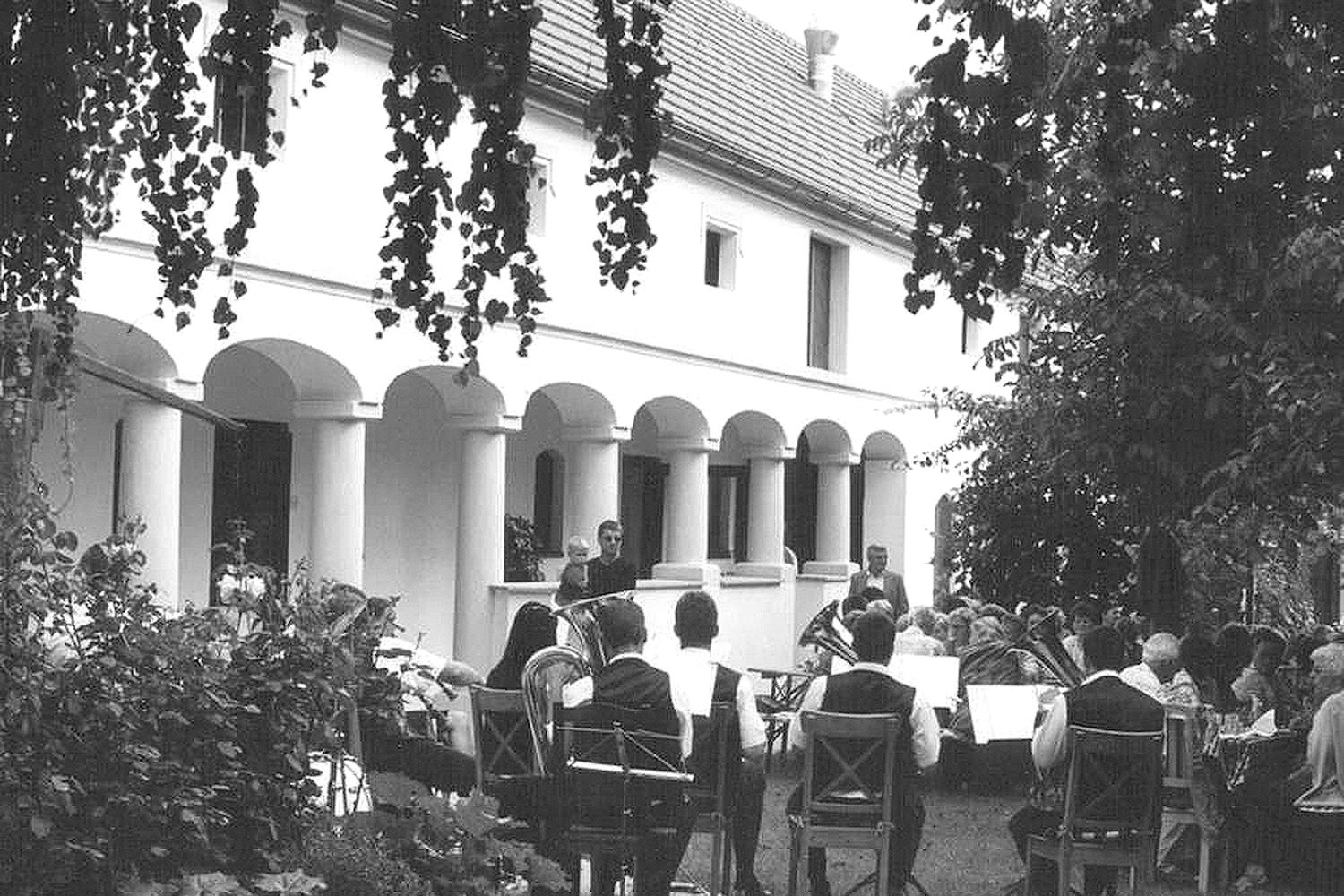 A group of people sit outside in front of a building with pillars while a brass band plays.