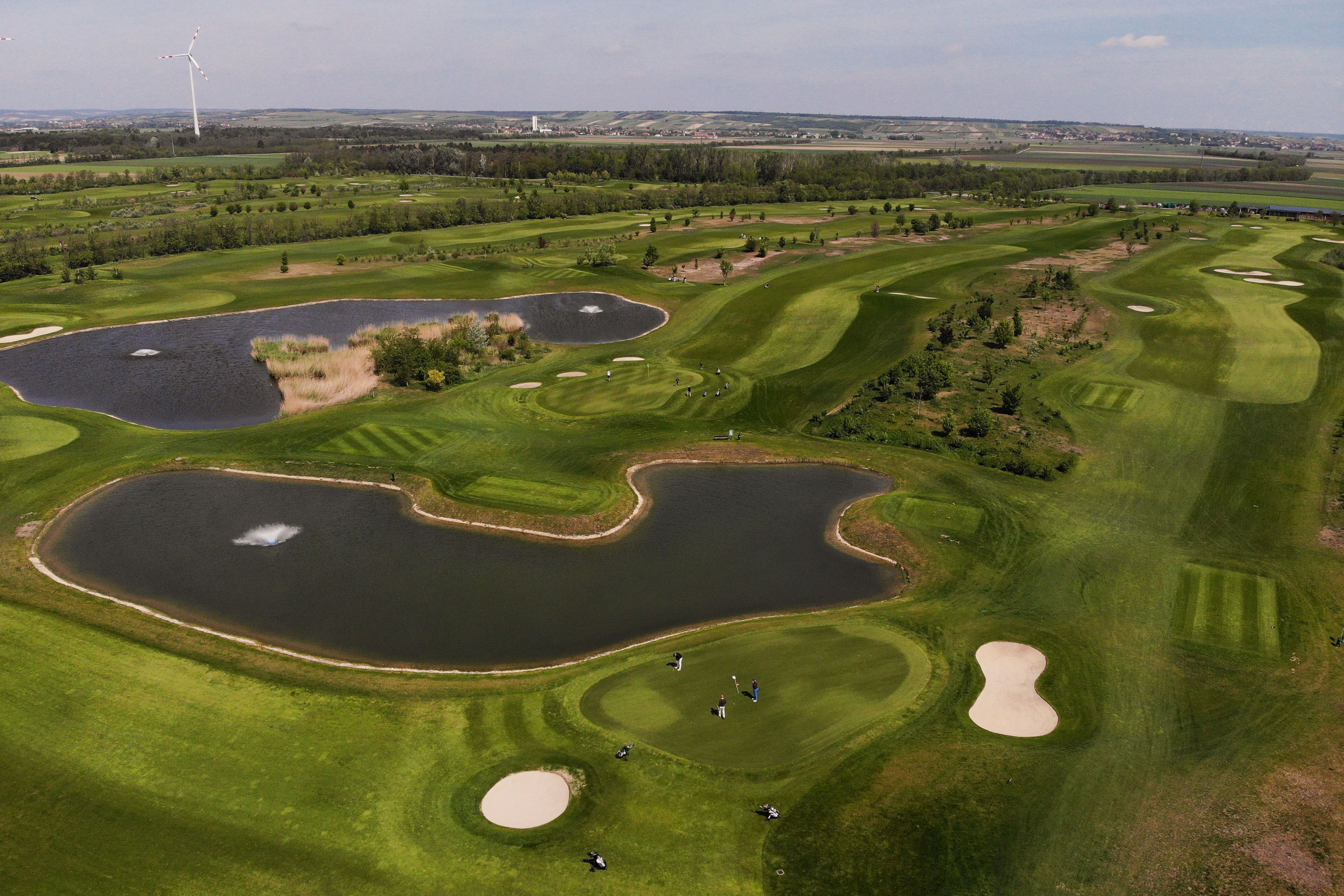 Aerial view of a golf course with ponds and wind turbine in the background.