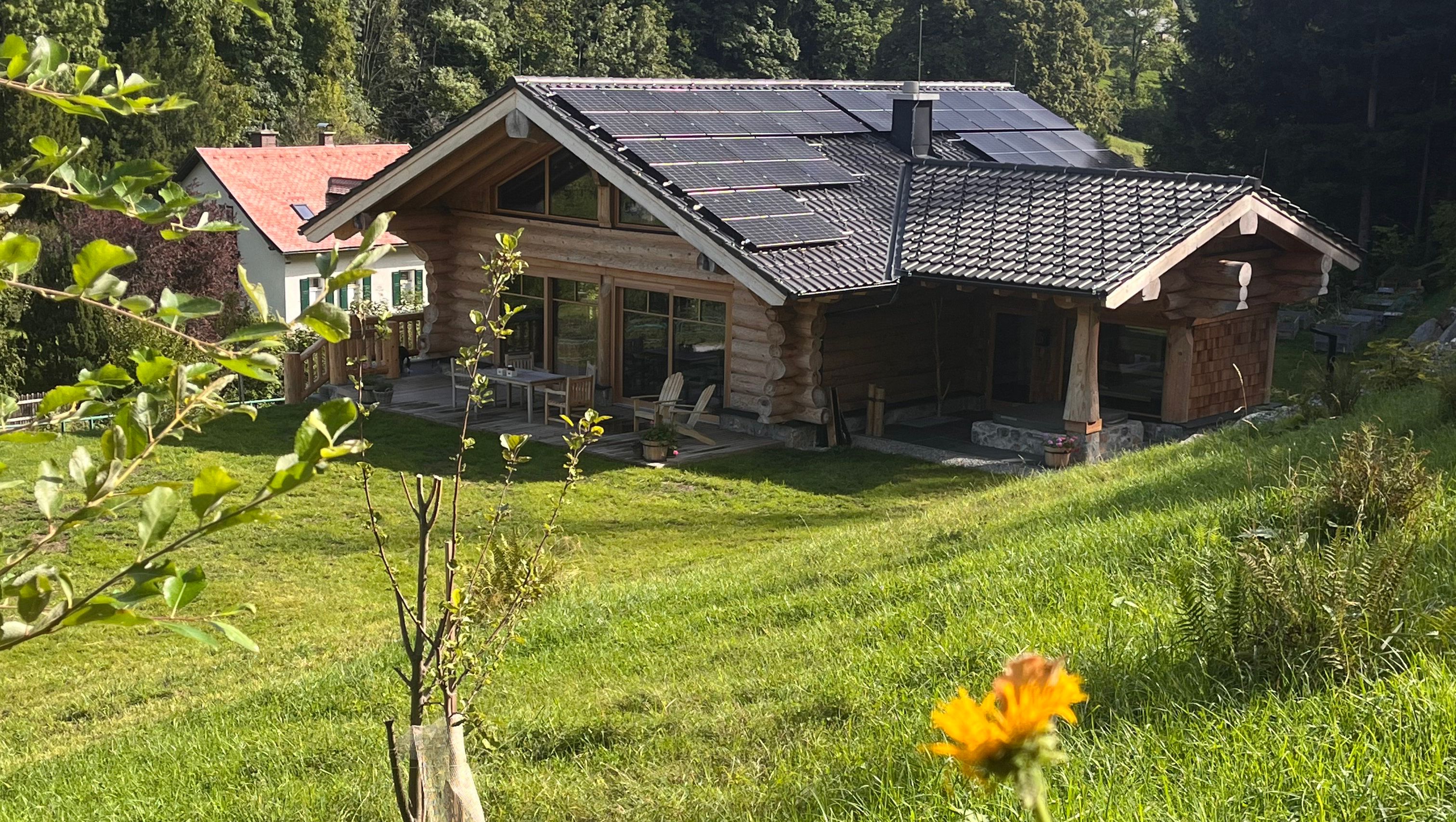 A wooden house with solar panels on the roof, surrounded by green landscape and trees.