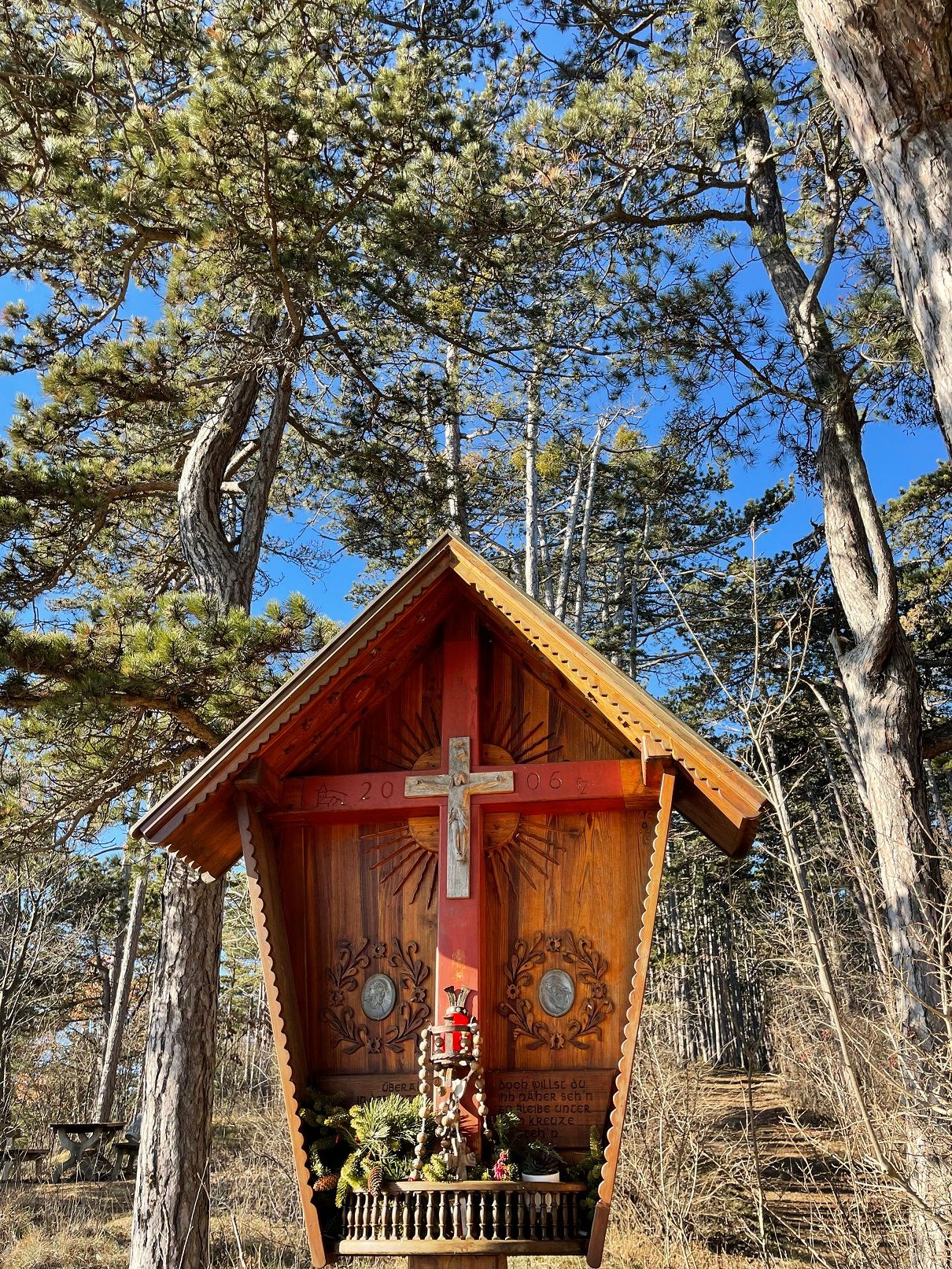 Wooden cross with roof in a forest, surrounded by trees and blue sky.