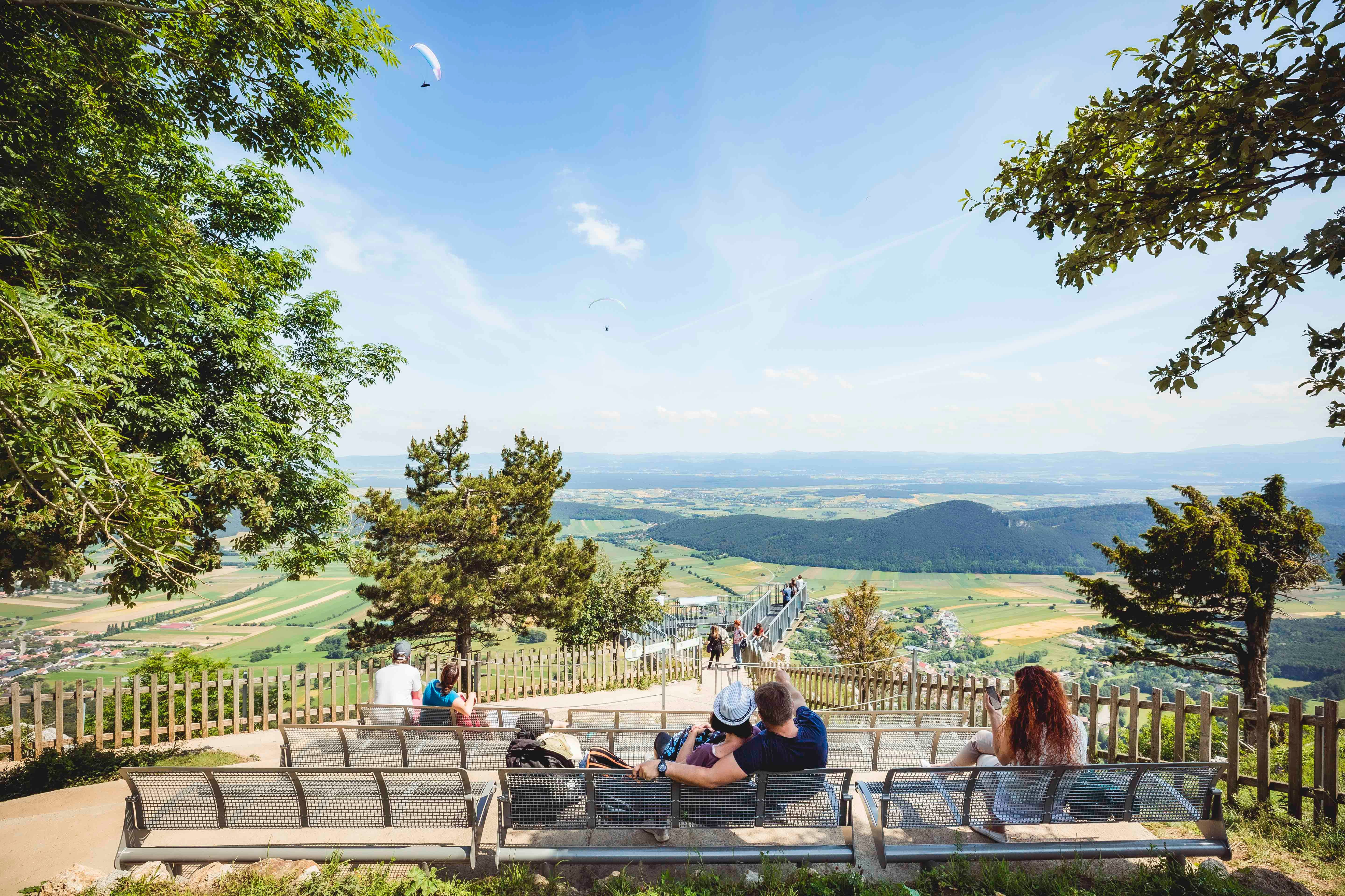 Viewing platform with a view of a vast landscape and paragliders in the sky.
