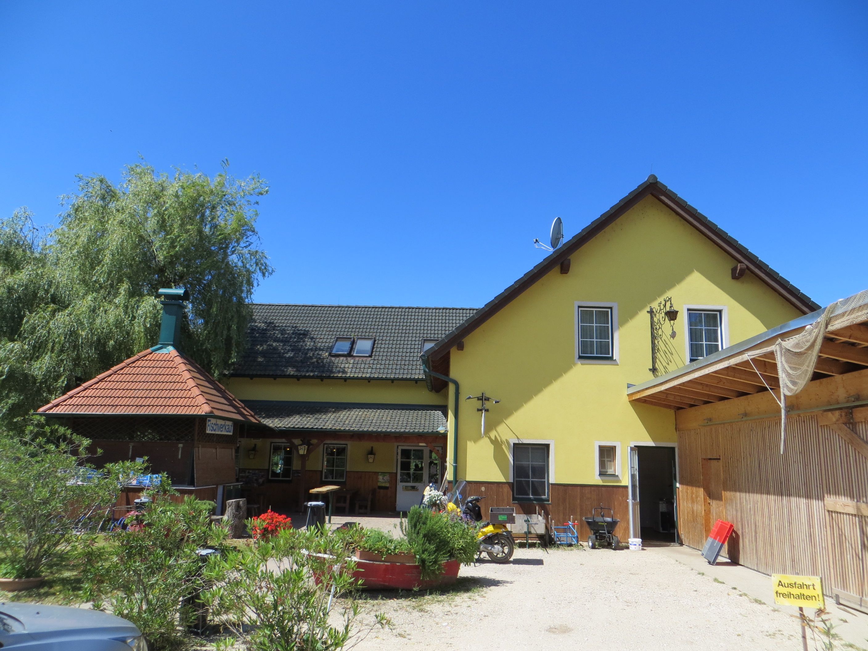 Yellow building with green roof and terrace, surrounded by plants, under a blue sky.