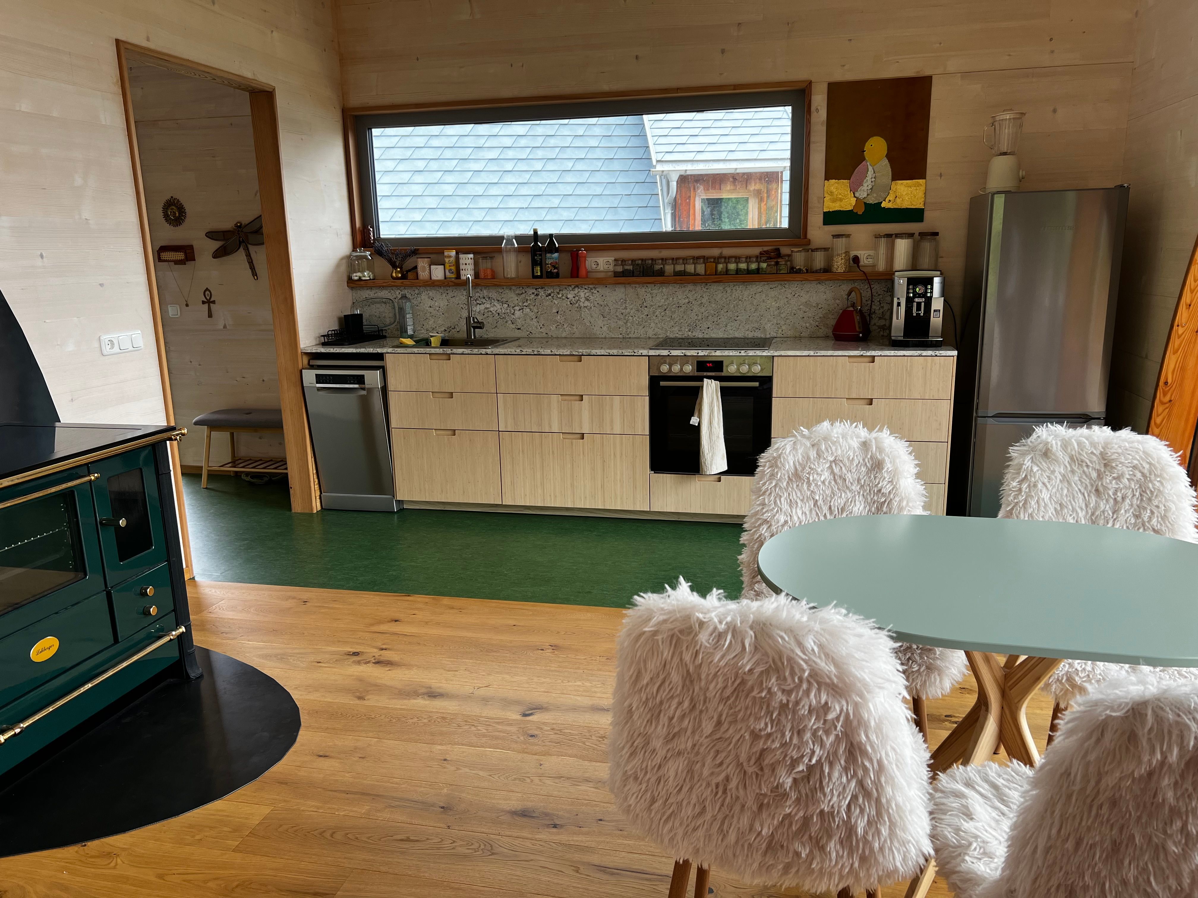 Modern kitchen with wood paneling, green stove, dining table with fluffy chairs and large window with view to the outside.