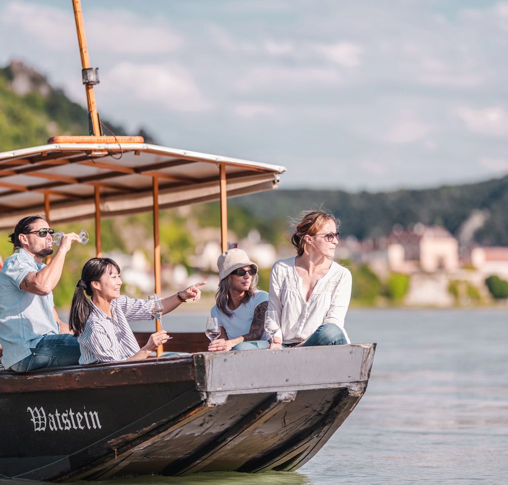 Eine Gruppe von Freunden genießt die sanfte Brise und die malerische Aussicht während einer entspannten Zillenfahrt auf dem Wasser. Die Sonne strahlt über die sanften Hügel, während sie mit einem Glas in der Hand die Schönheit der Natur bewundern.