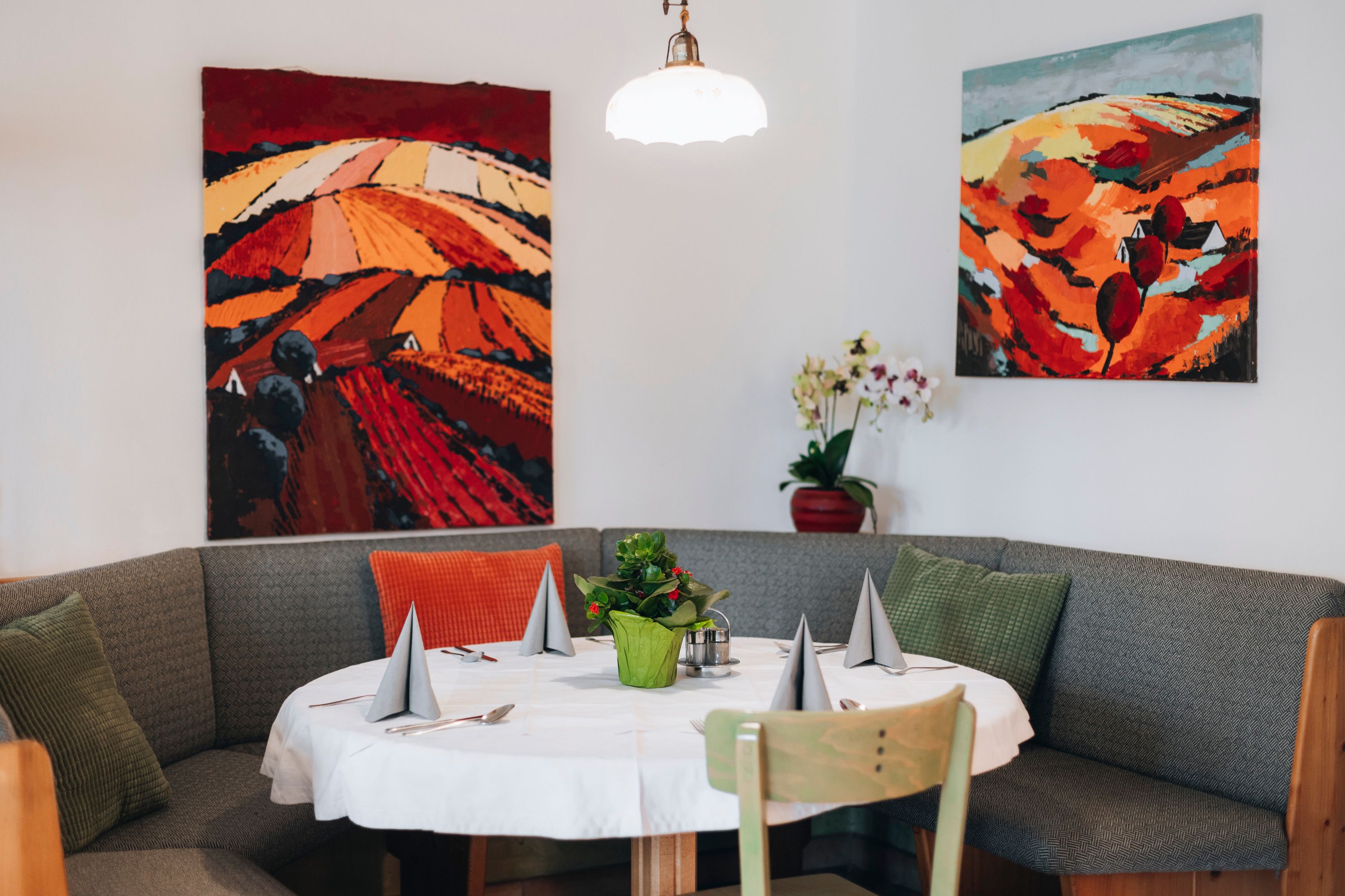 Cozy seating area with table, decorated with grey napkins and a plant, surrounded by colorful landscape paintings on the wall.