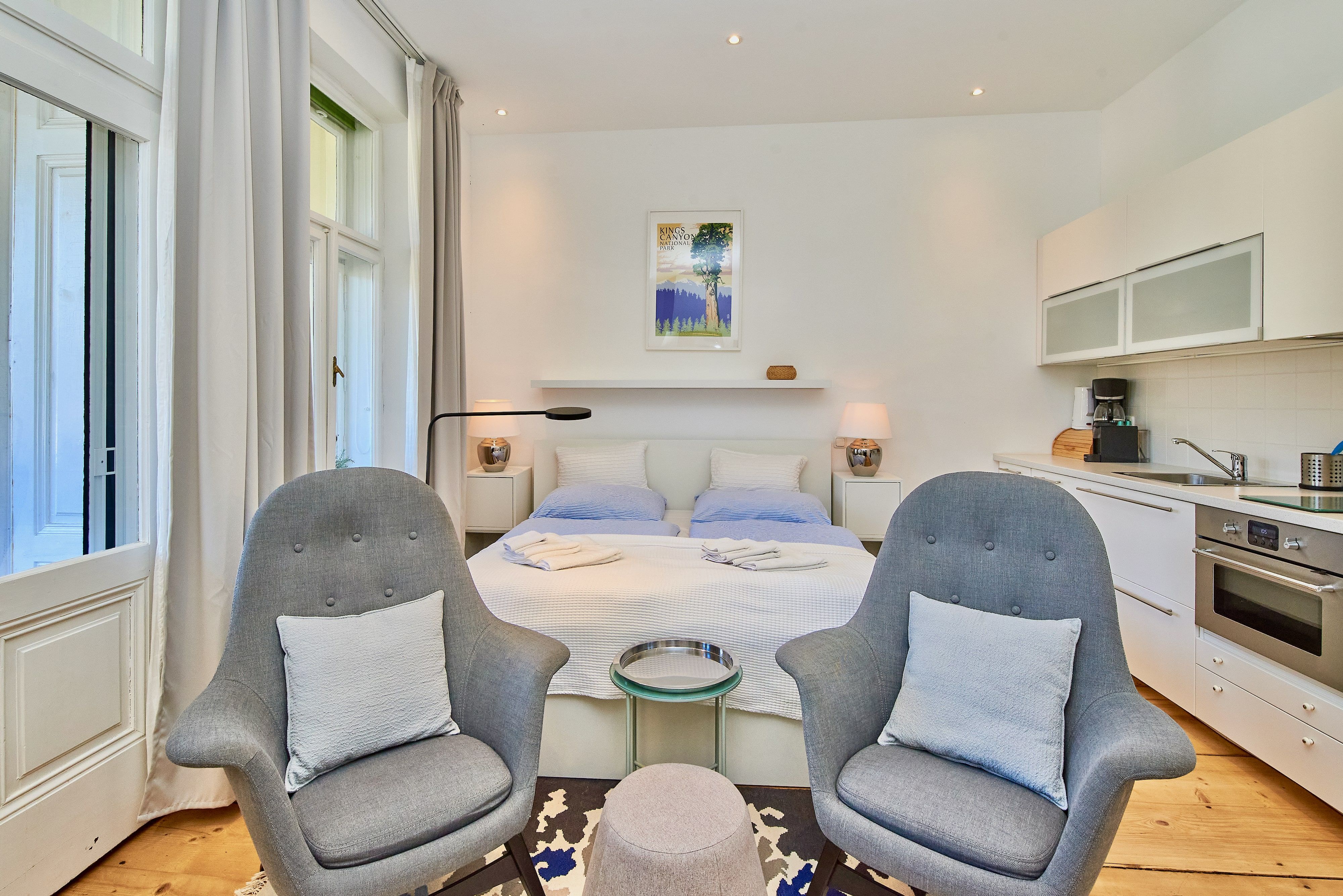 Modern studio apartment with double bed, two gray armchairs, kitchenette and large windows.