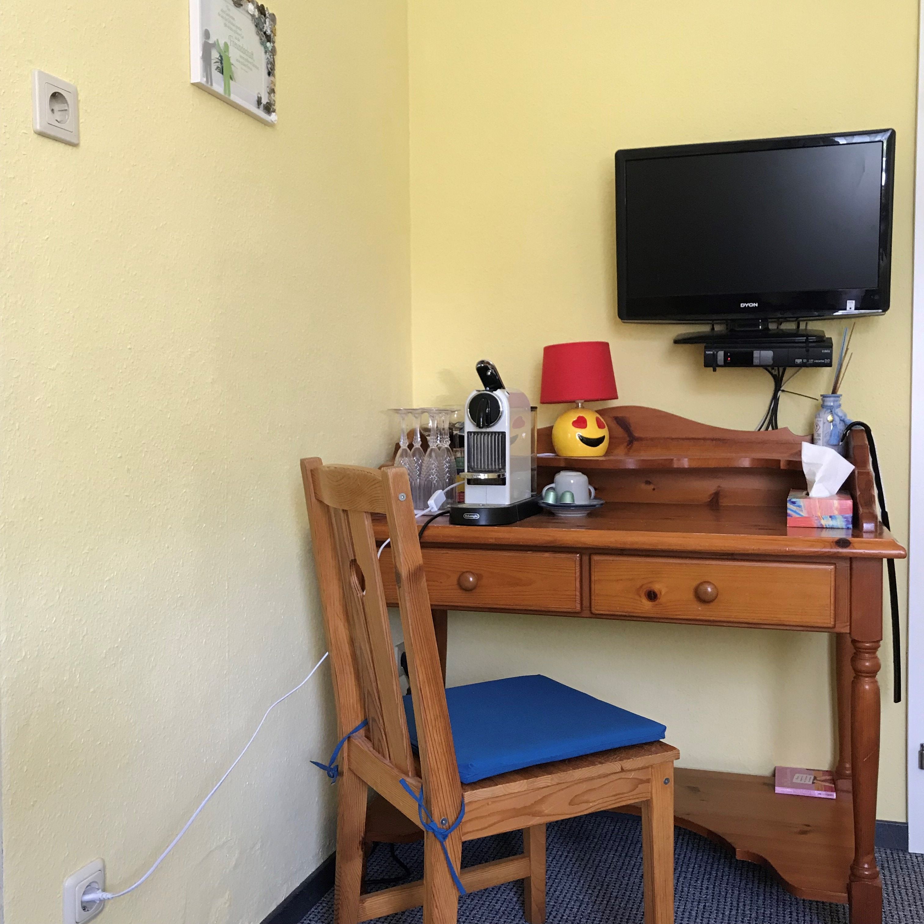 A small wooden table with a coffee machine, glasses, a lamp and a TV on the wall.