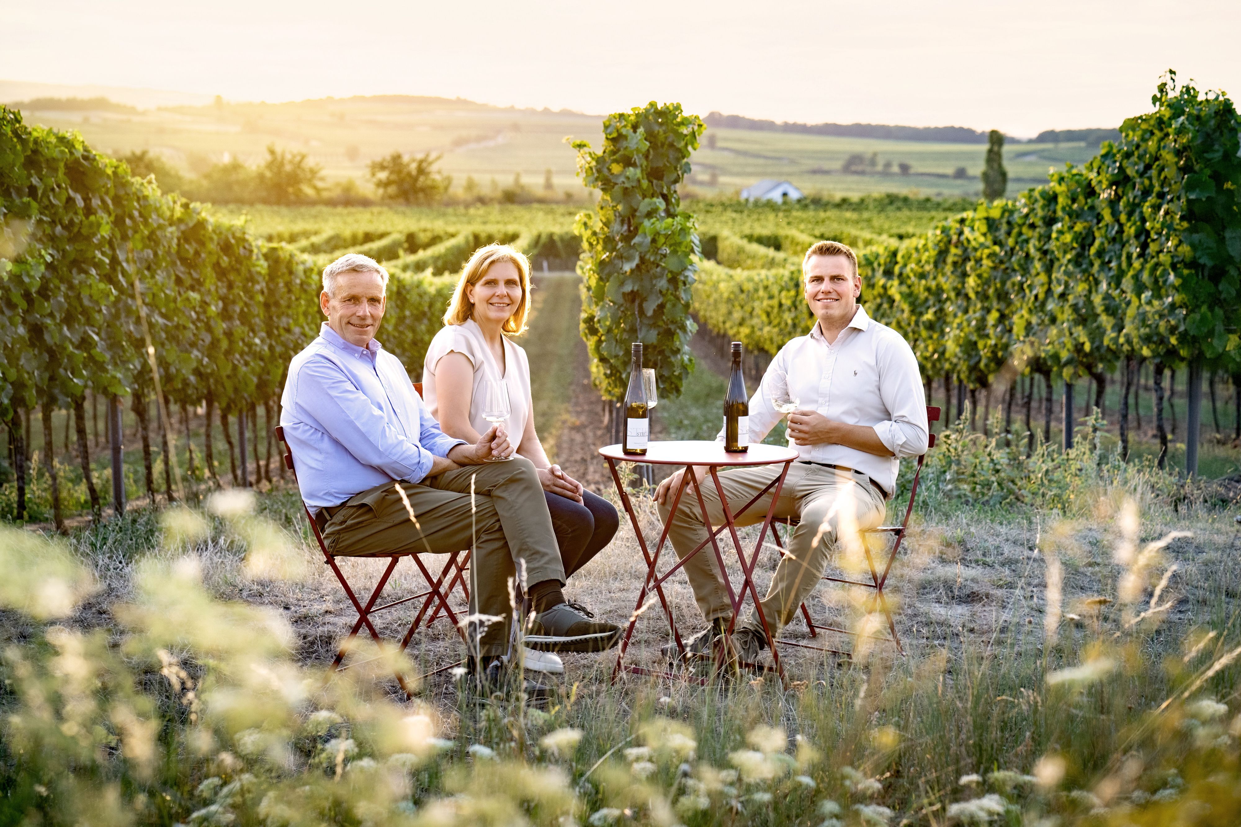 Three people sit at a table in the vineyard with wine glasses and bottles.