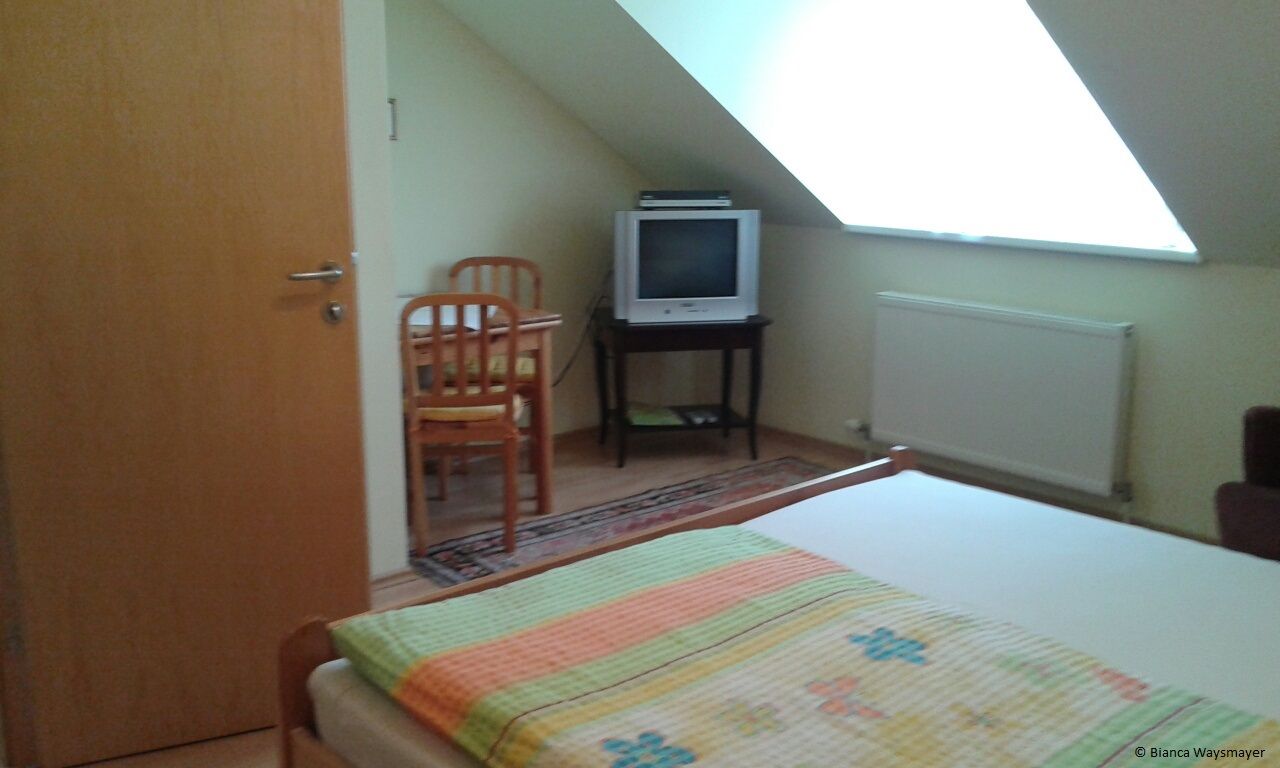 A simply furnished room with a bed, TV and table under a sloping roof.