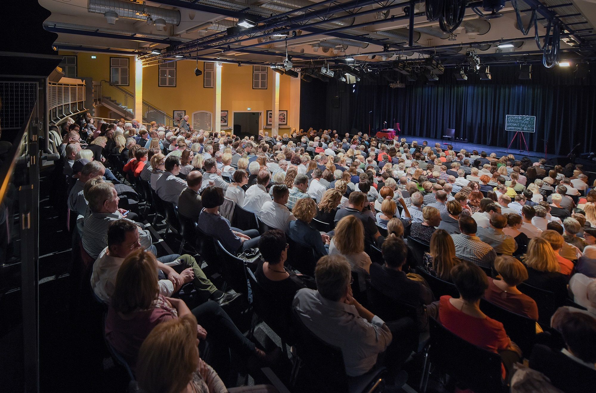 A packed theater hall with an audience looking onto a stage.