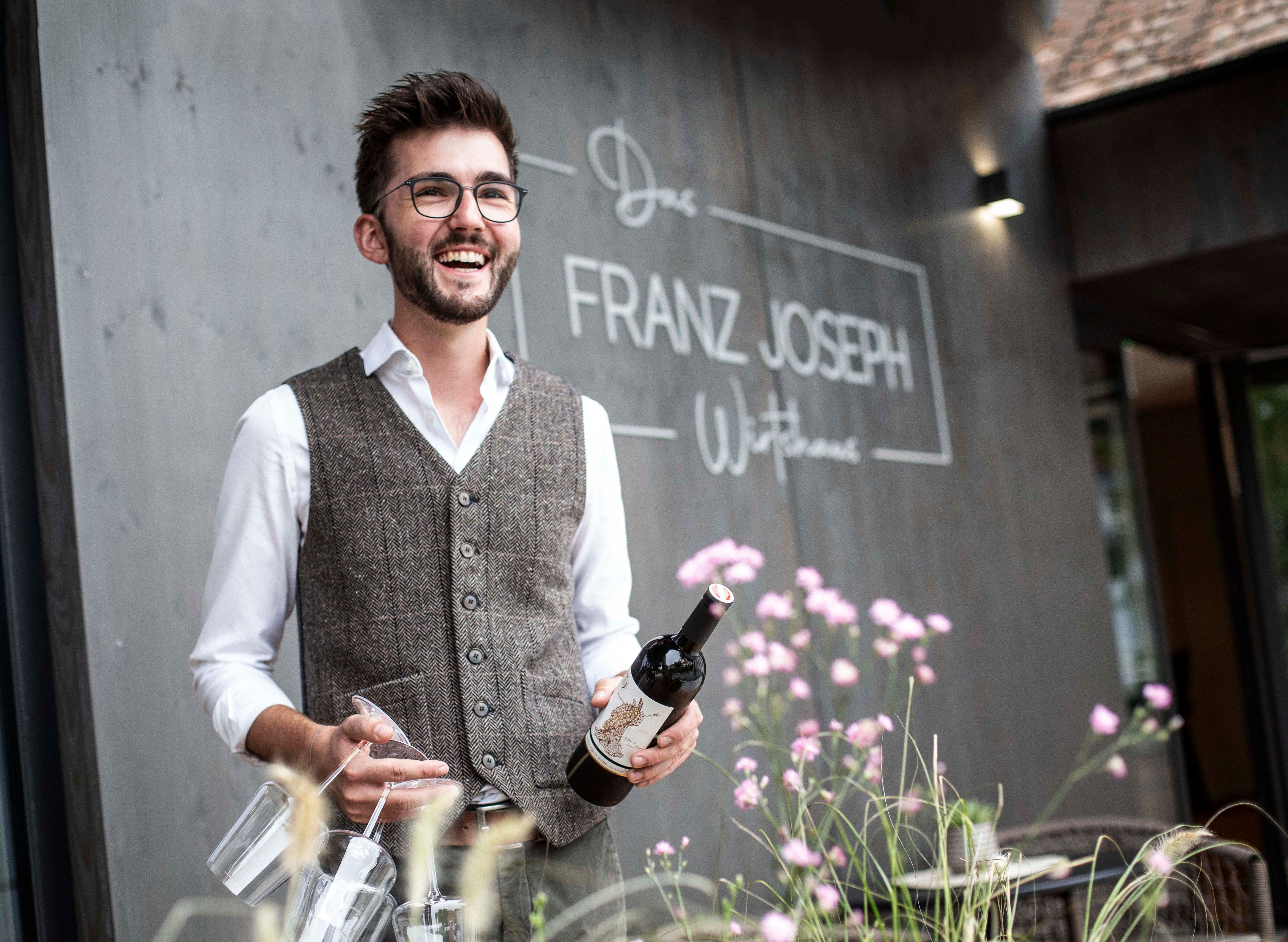 Man with glasses and vest holding wine bottle in front of restaurant 'Franz Joseph'.
