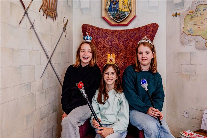 Three children with crowns and sceptres sit on a throne in a fairytale room.