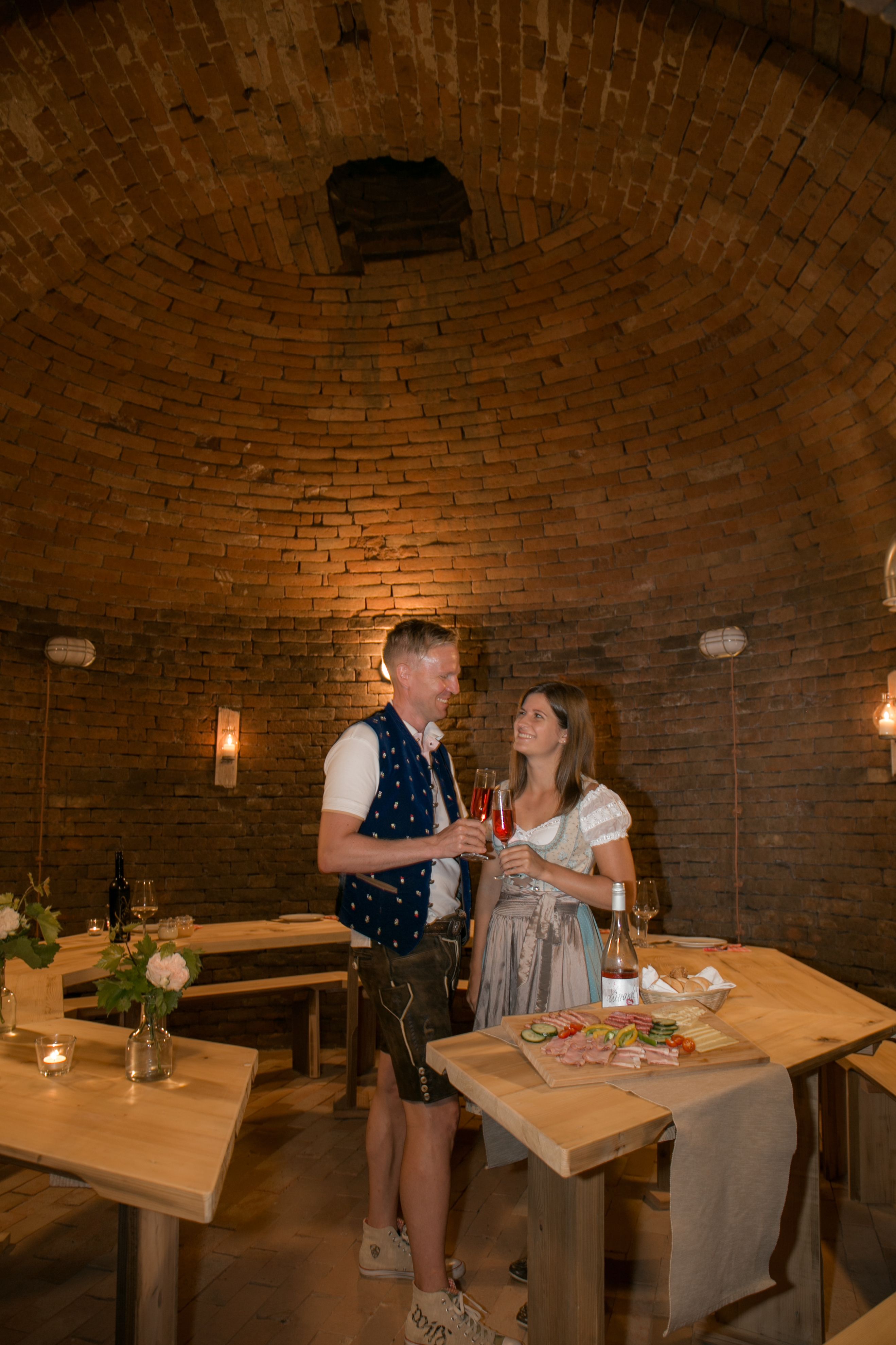 A couple in traditional dress toast with drinks in a rustic, vaulted room.
