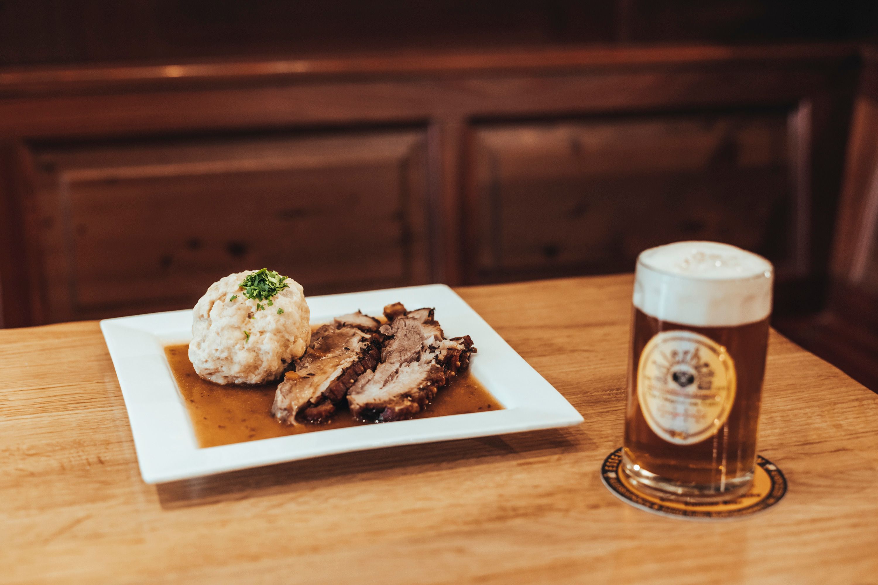 A plate of roast pork and dumplings next to a glass of beer on a wooden table.