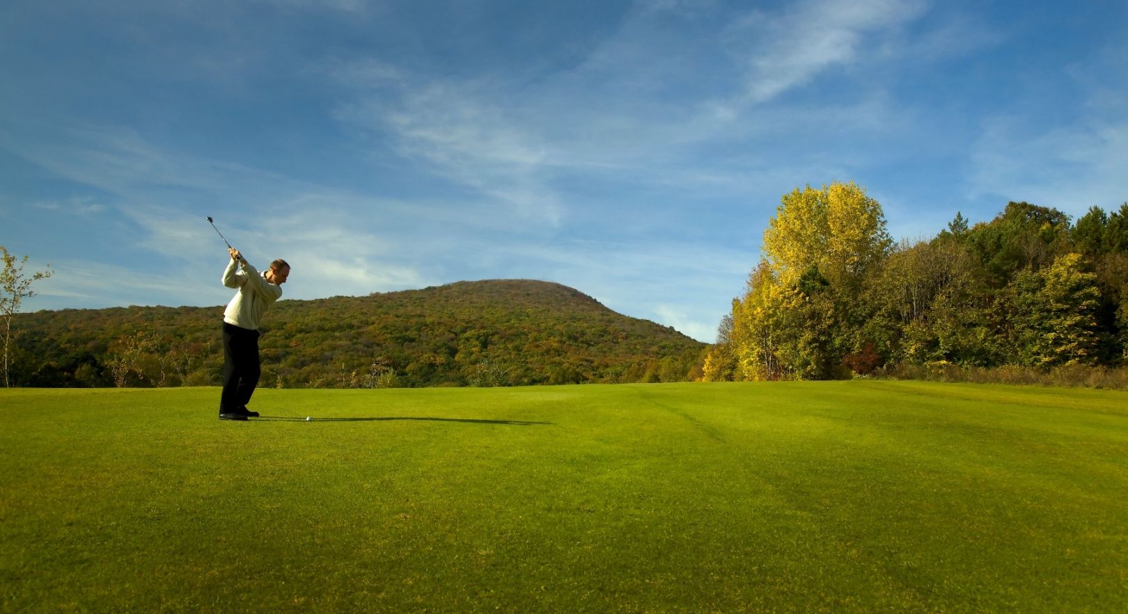 Golfer hits ball on a green golf course with wooded hills in the background.
