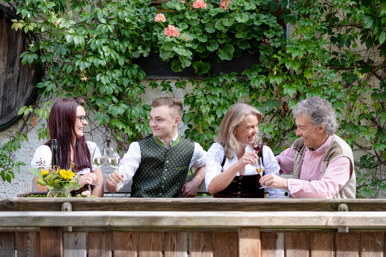 Four people in traditional dress clink glasses of wine outside in front of a wall covered in plants.