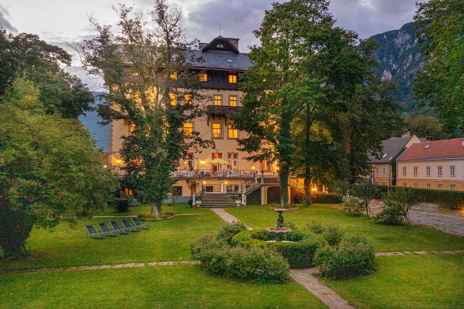 An illuminated building with a garden and sun loungers at dusk.