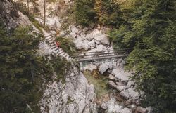 Zwei Wanderinnen überqueren eine Holzbrücke über einen felsigen Bachlauf in einer bewaldeten Schlucht