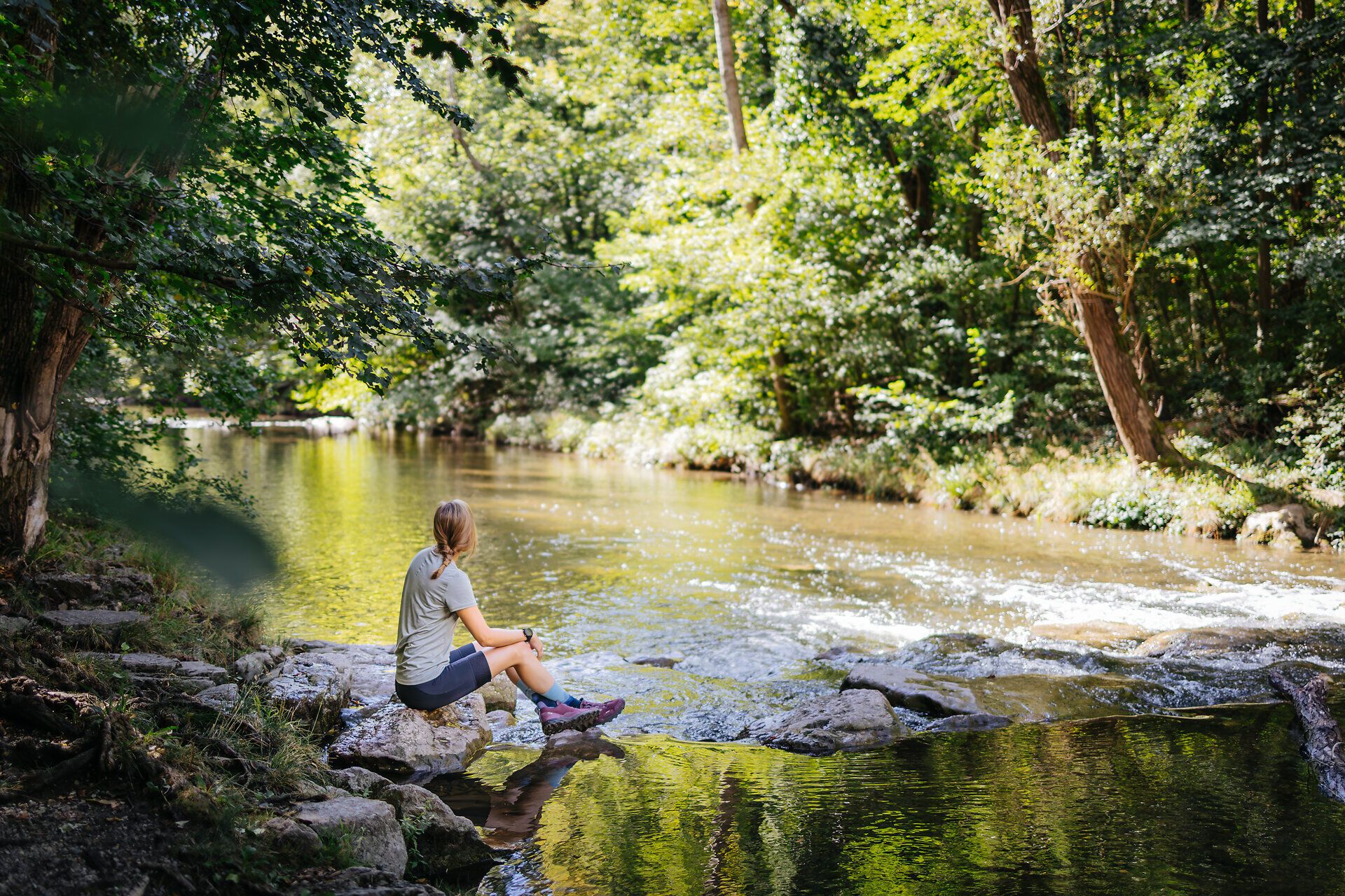 Ein sanfter Fluss schlängelt sich durch die üppige, grüne Landschaft, während die Sonne durch das Blätterdach der Bäume strahlt. Die ruhige Atmosphäre lädt dazu ein, die Seele baumeln zu lassen und die Schönheit der Natur zu genießen.