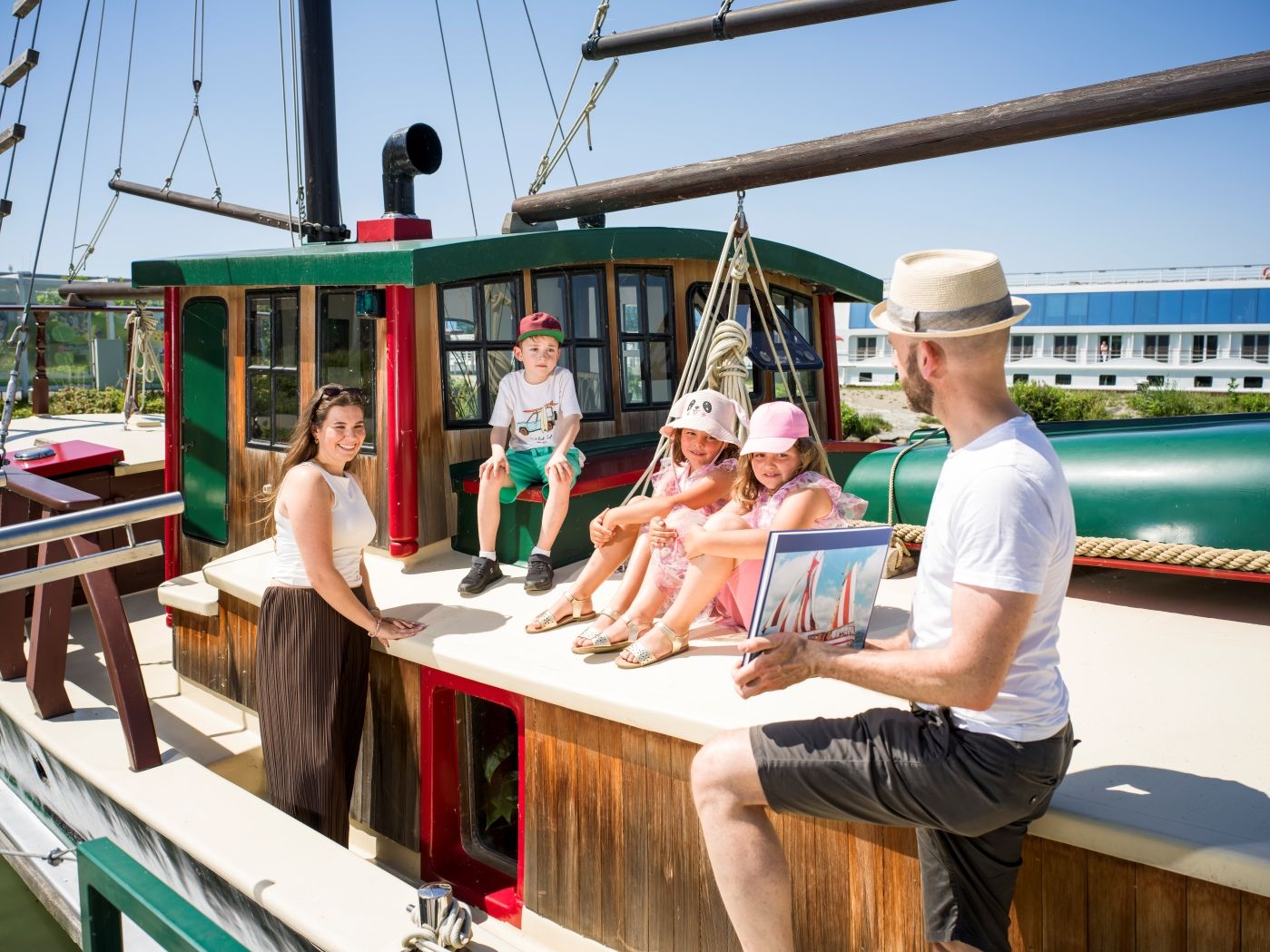 Children are sitting on the deck of the historic Hundertwasser ship; a woman is standing next to them, and the tour guide is showing photos of the ship