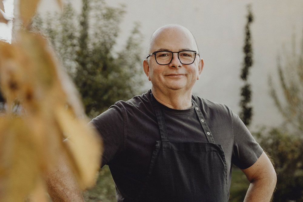 Man with glasses and apron smiling outdoors, blurred background.