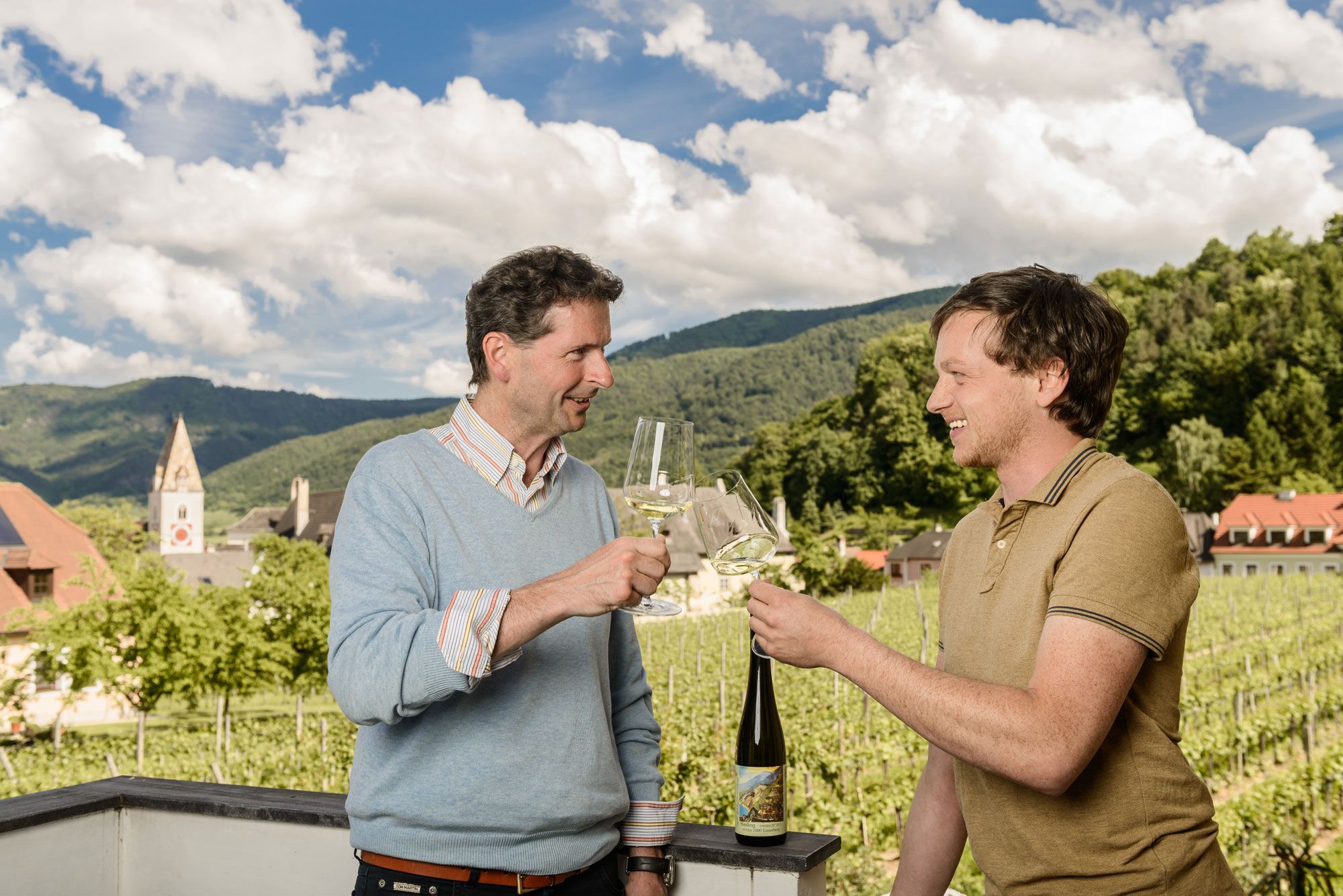 Two men clink glasses of wine, with vineyards and a church tower in the background.