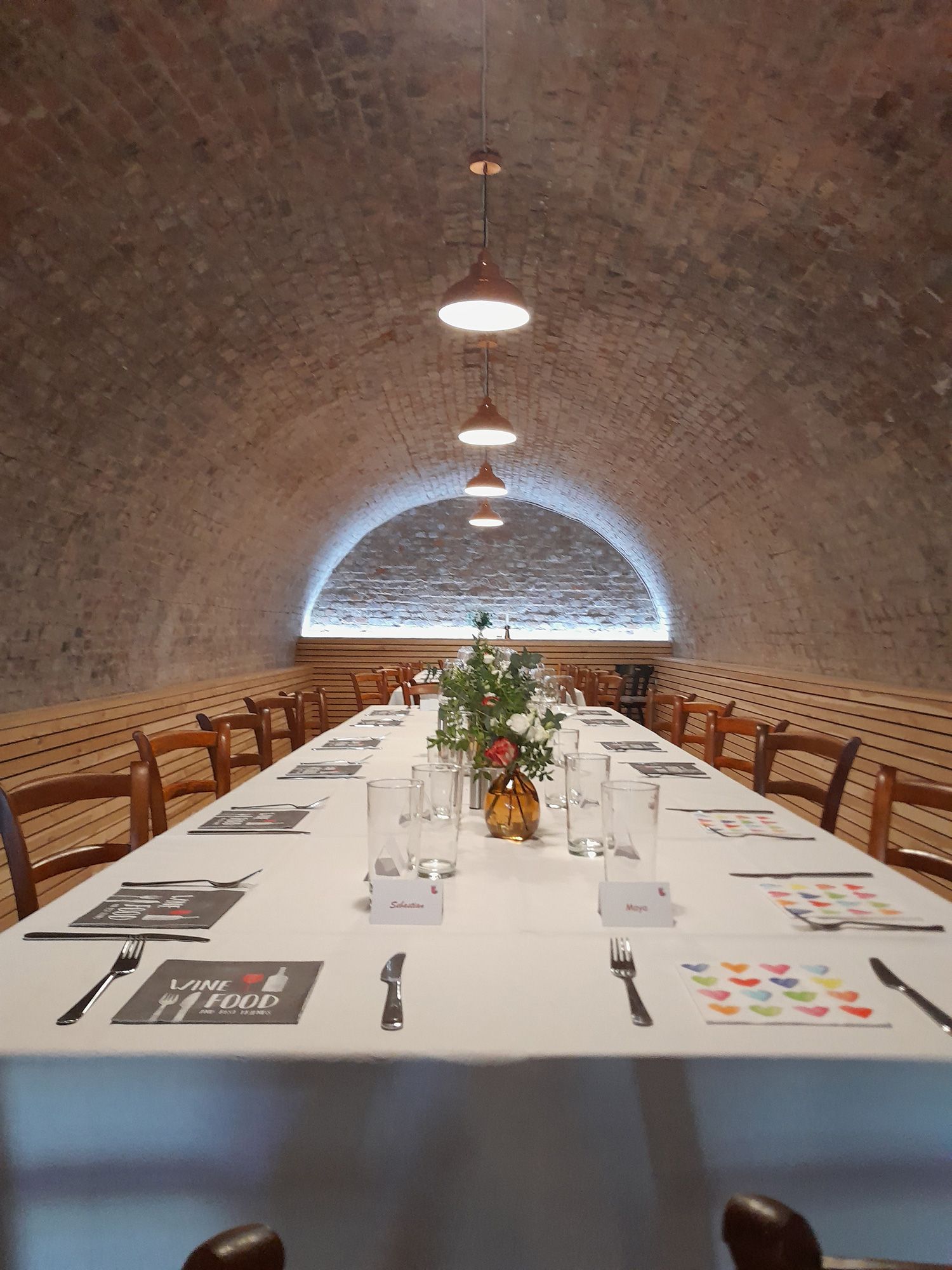 Long table in a vaulted cellar, set with white tablecloths, glasses and flowers.