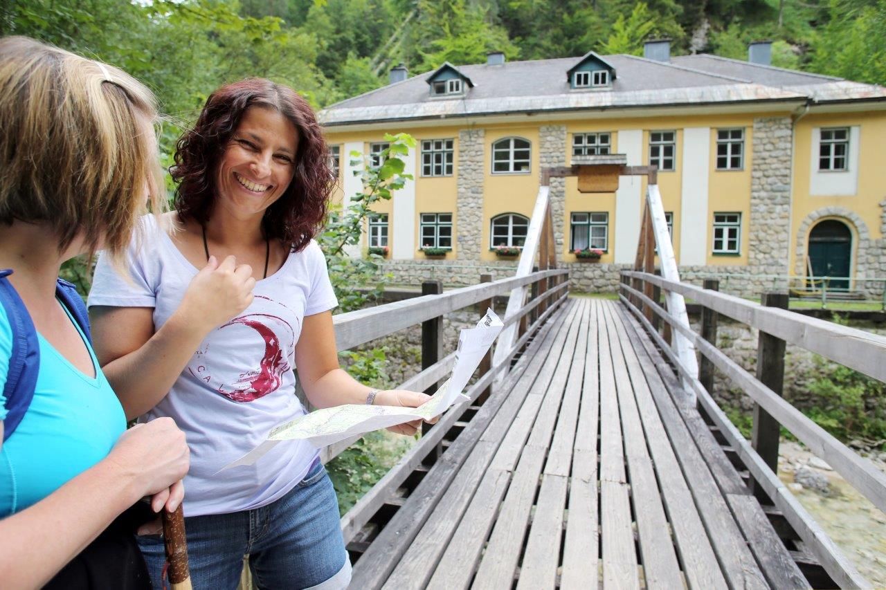 Two women stand on a wooden bridge in front of a yellow building in Wienerbruck, one holding a map.